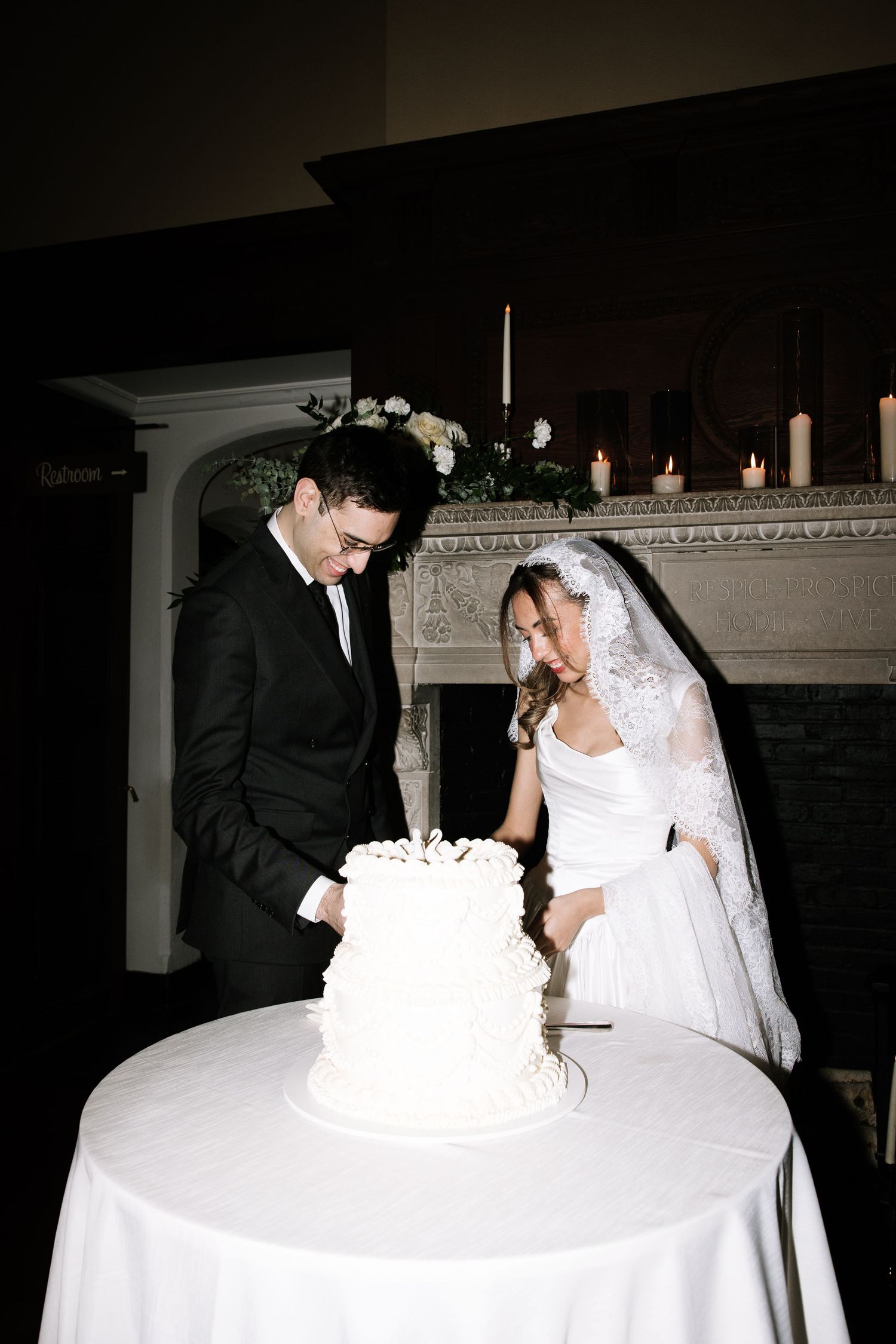 Bride and groom cutting white wedding cake at elegant reception with candlelit fireplace backdrop.