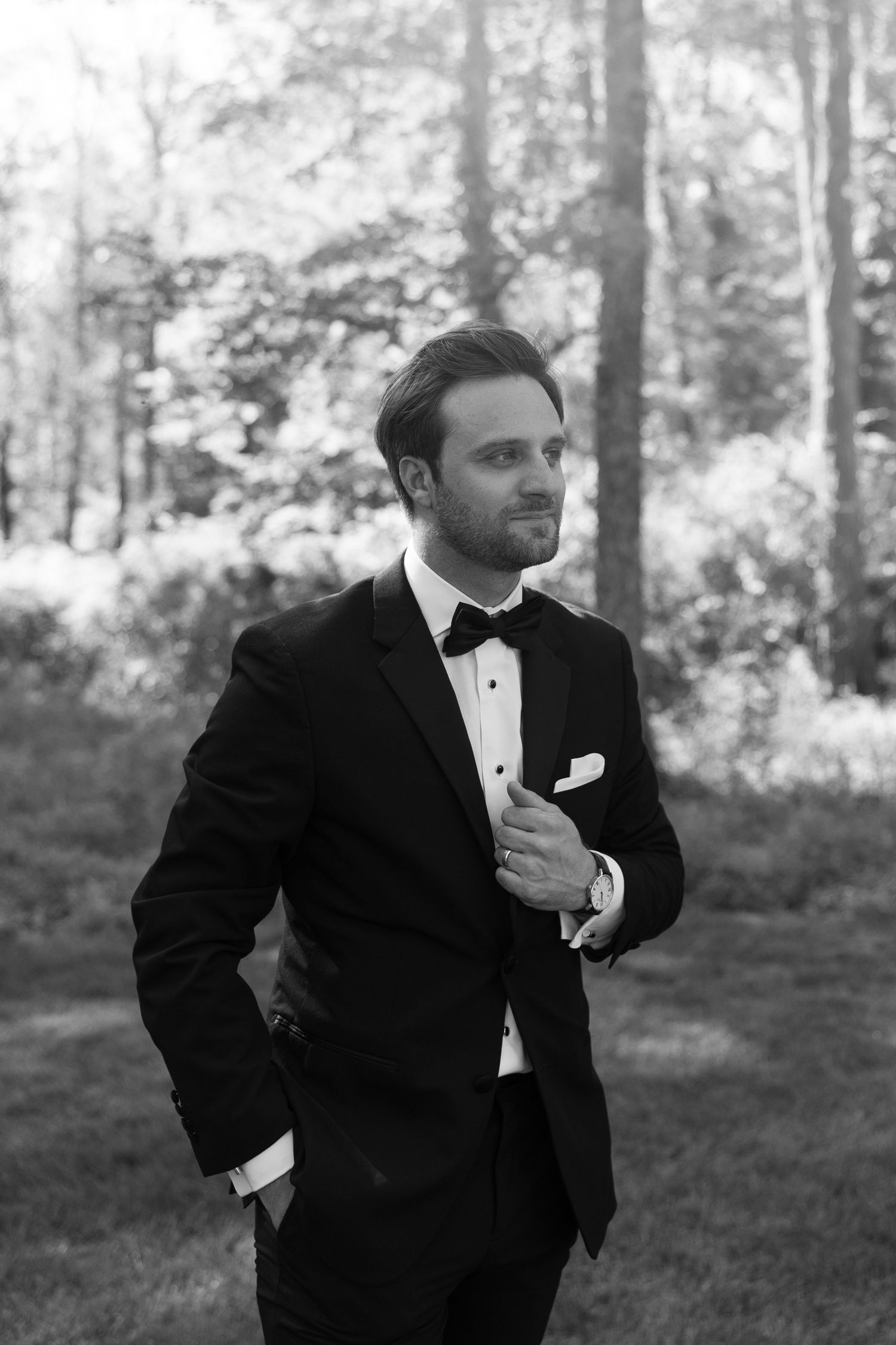 Black and white photo of a well-dressed man in a tuxedo with bow tie standing outdoors among trees.