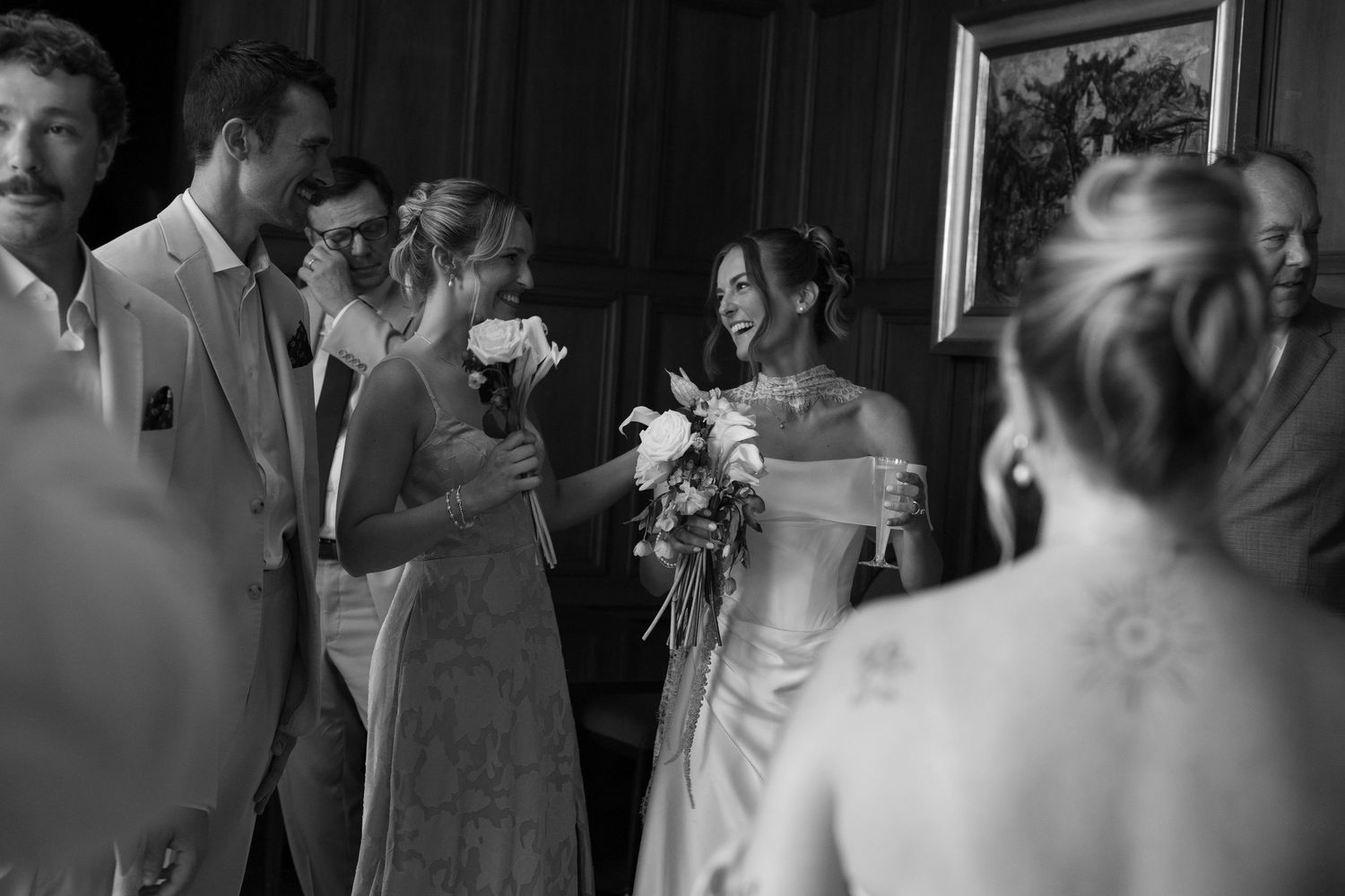 Black and white wedding photo of smiling bride holding bouquet greeting guests indoors.