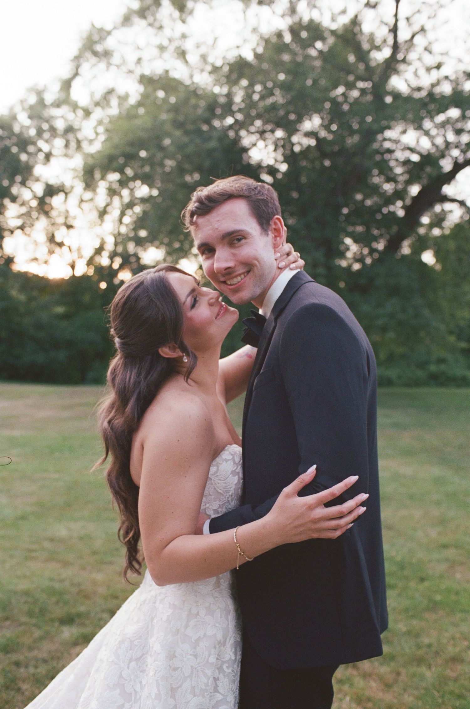 A bride in a lace gown embraces a groom in a navy tuxedo outdoors at golden hour.