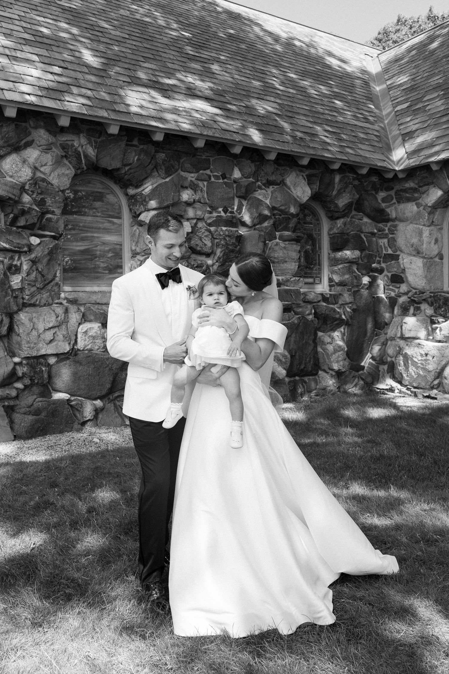 Black and white photo of a bride and groom in formal wear holding their baby outside a stone chapel.