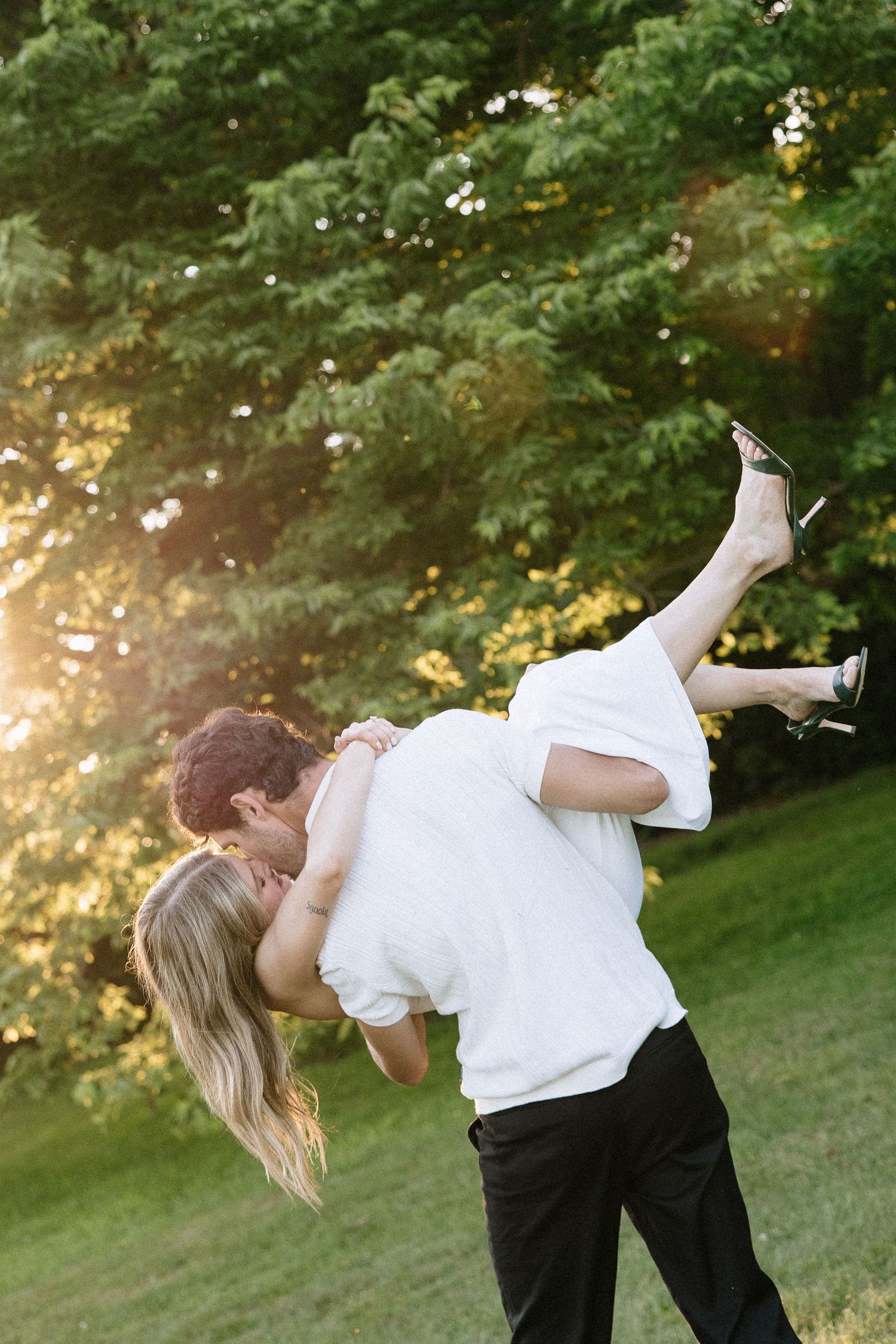 Couple sharing a romantic kiss at golden hour in a lush green park, man lifting woman in white dress.
