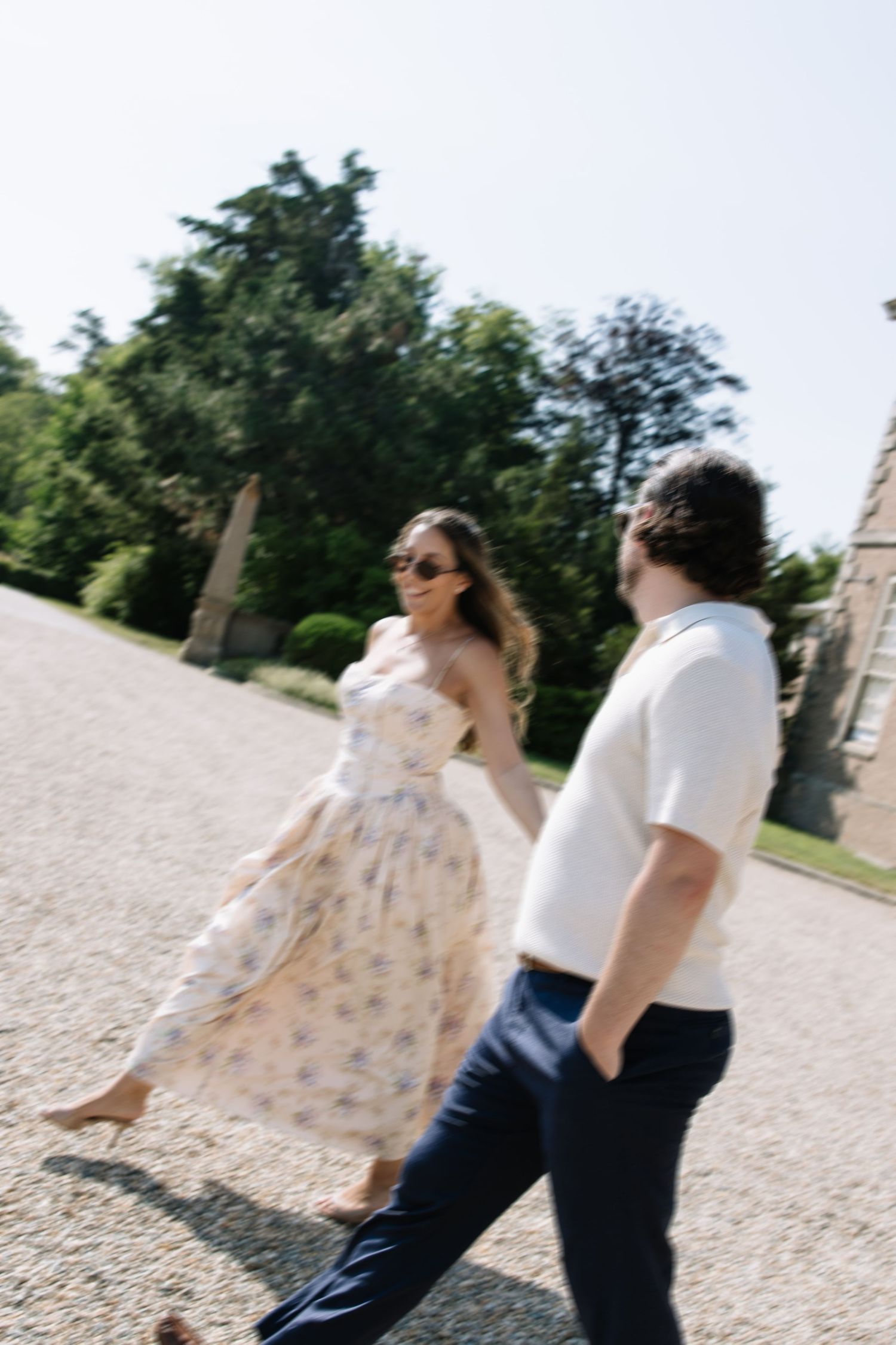 Couple walking outdoors on a sunny day, woman in floral dress and sunglasses, man in white shirt and navy pants.