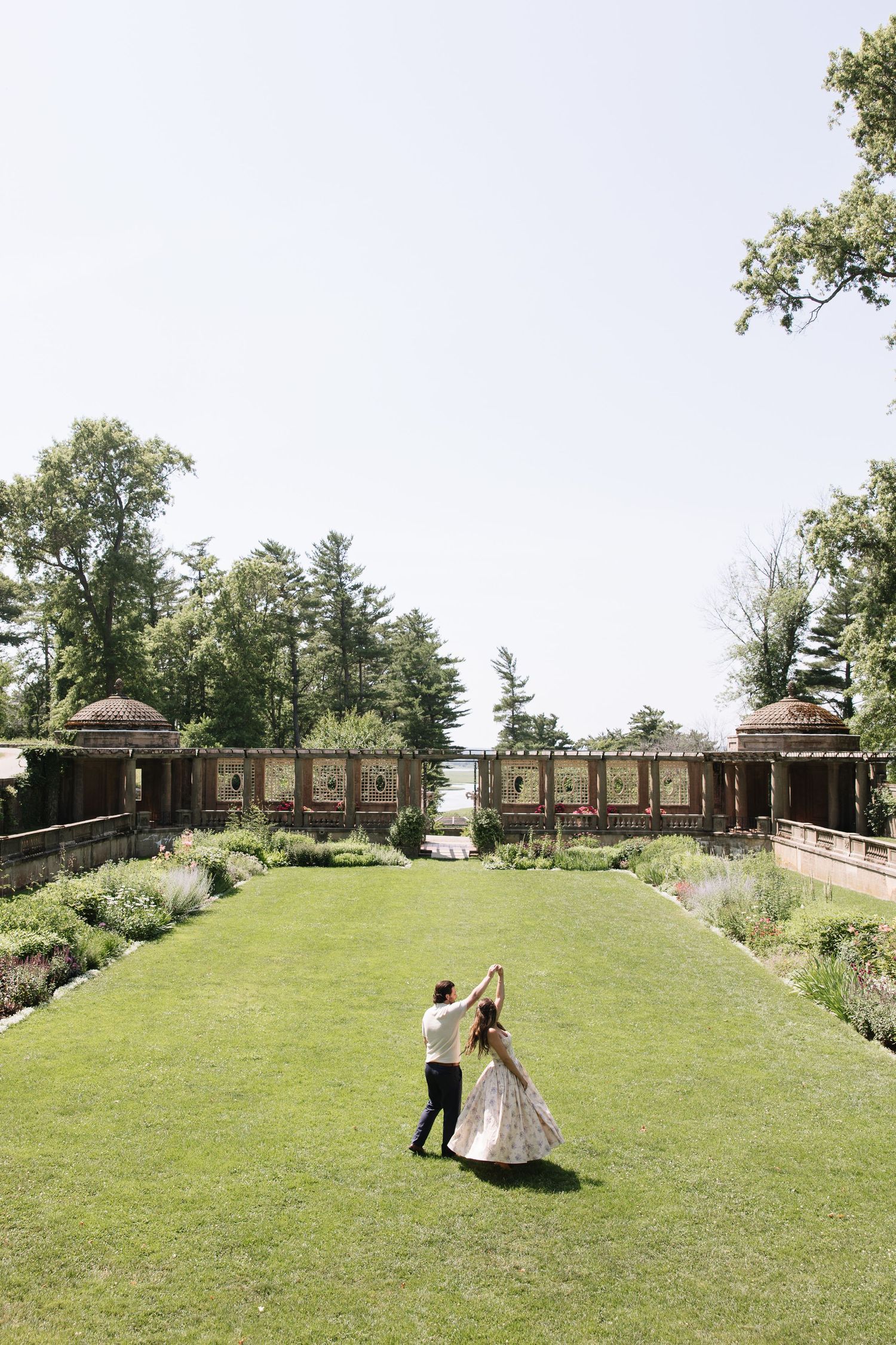 Bride and groom dancing on lush green lawn of elegant garden venue surrounded by trees on sunny wedding day.