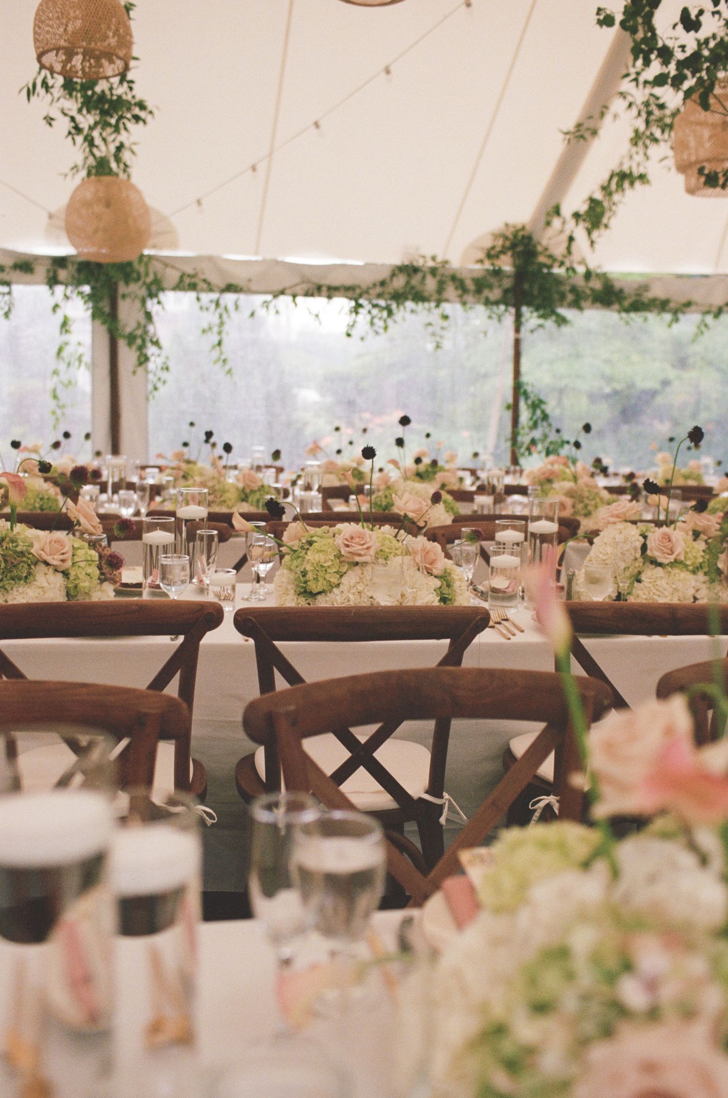 Elegant outdoor wedding reception tent with wooden cross-back chairs, floral centerpieces, and lush greenery garlands.