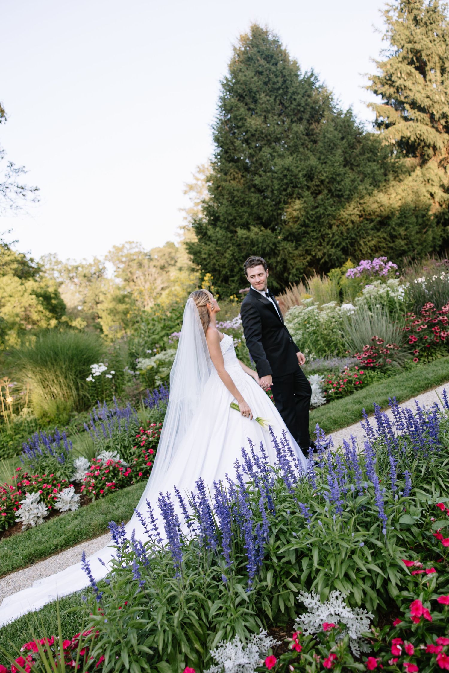 Wedding couple poses in a garden with purple lupines and pink flowers blooming along a scenic path.