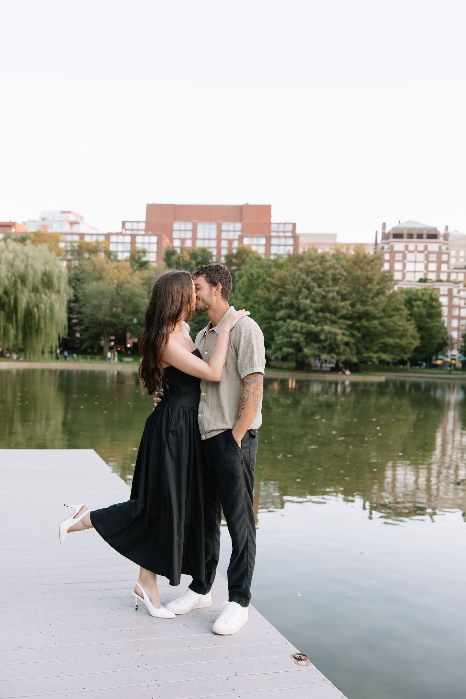 A couple shares a romantic moment by a pond with city buildings in the background.