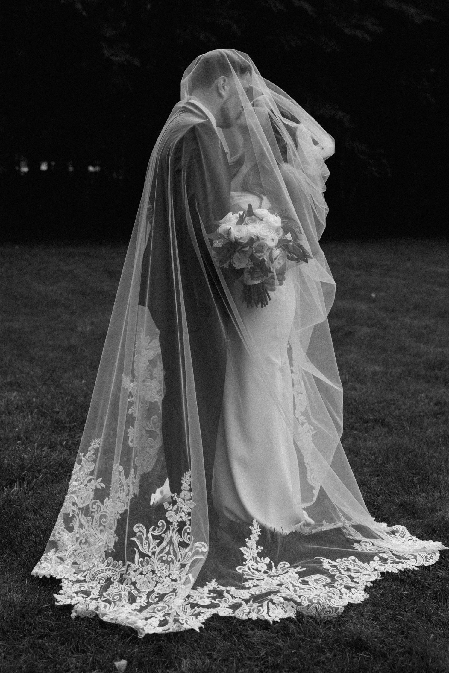 Detailed lace trim on wedding veil spreads across grass in black and white.