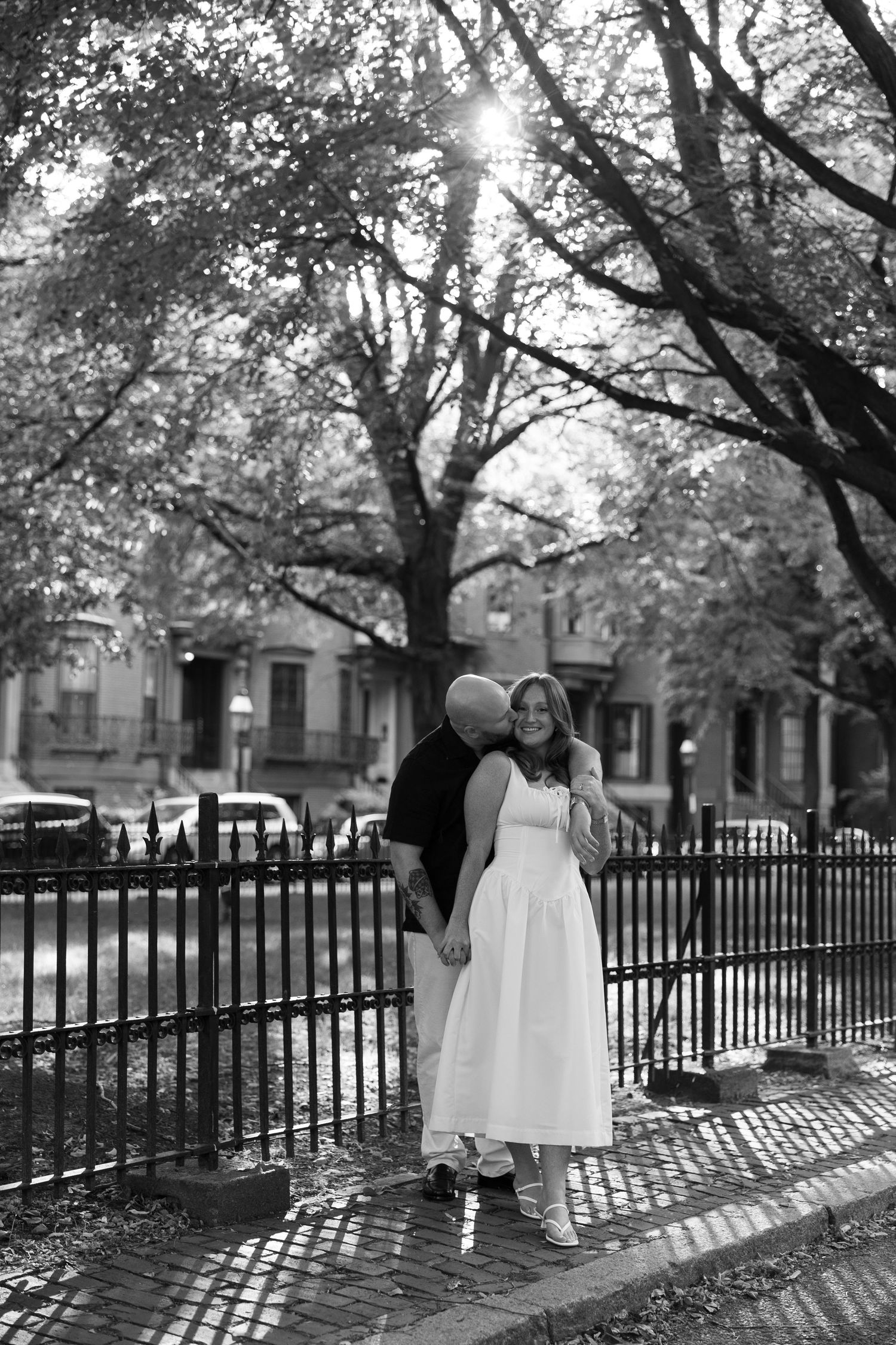 Black and white photo of couple standing together by iron fence under tree canopy.