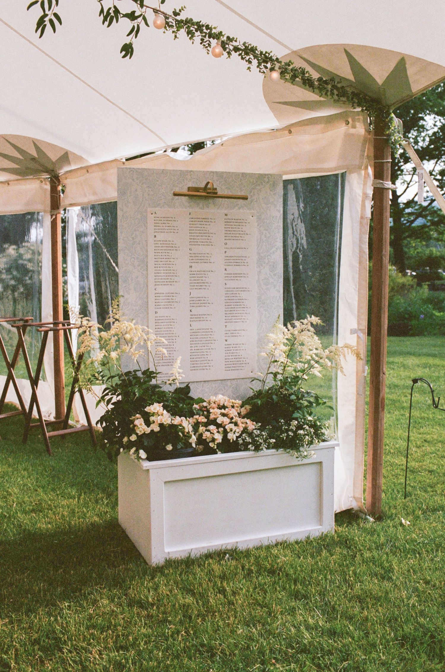 Elegant outdoor wedding seating chart display with white planter box filled with flowers under a tent.