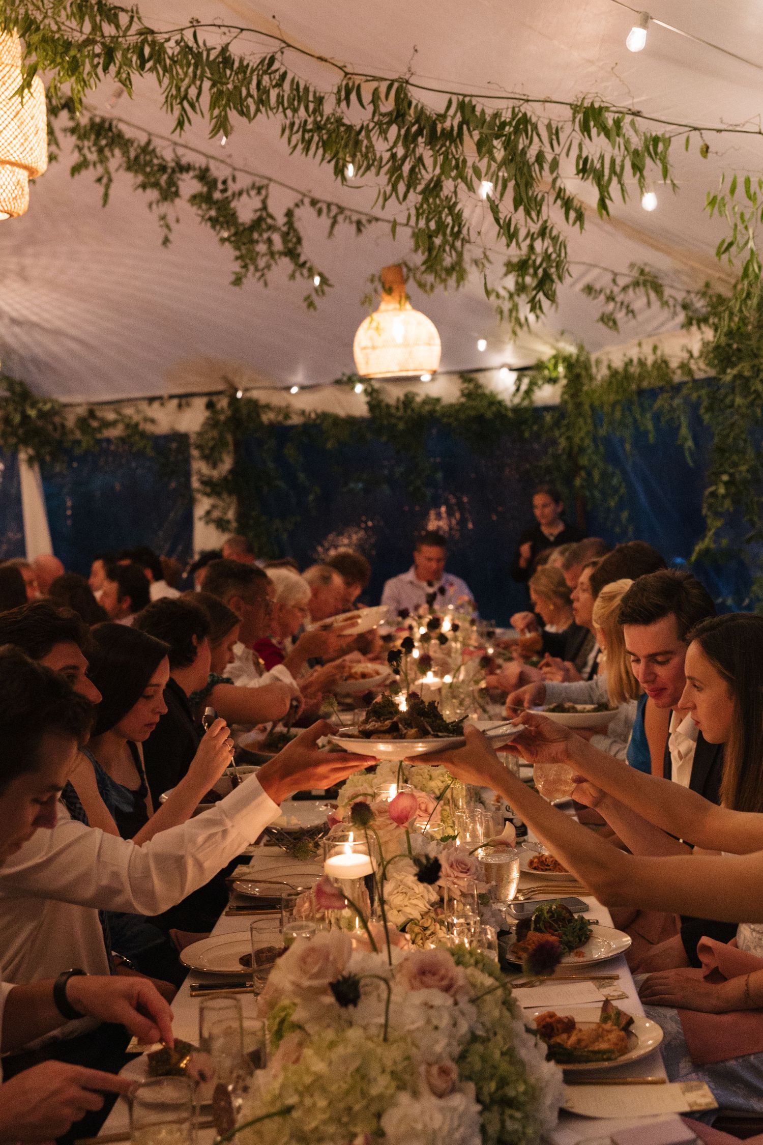 Intimate candlelit wedding reception dinner under tent with guests seated at long tables adorned with floral centerpieces and string lights.