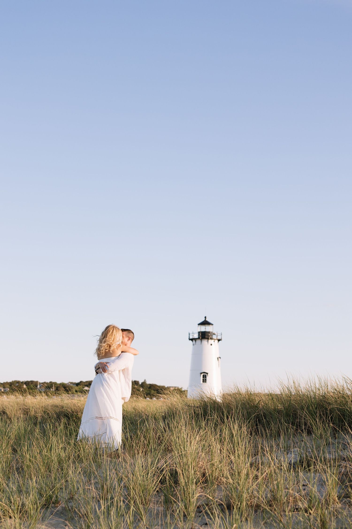 Couple embracing in tall grass near a white lighthouse on a clear sunny day at the beach.