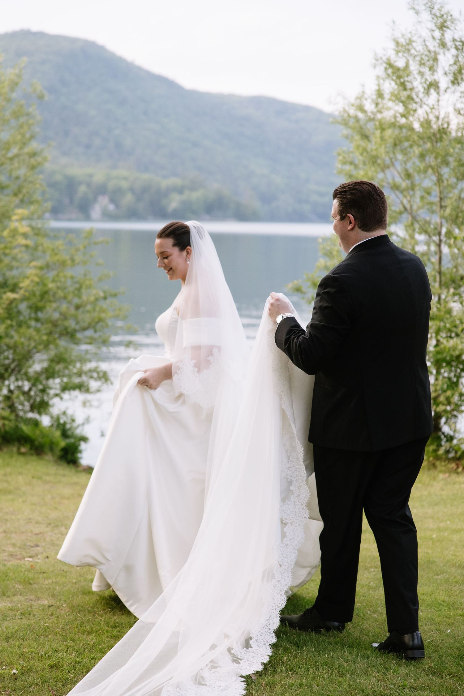 Groom adjusts bride's lace-trimmed cathedral veil outdoors, with a serene lake and green mountains in the background.