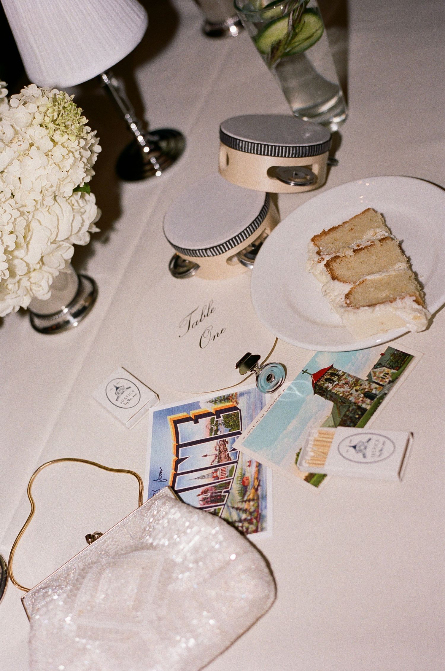 Elegant wedding table with cake slice, postcards, sparkly clutch, and floral centerpiece.