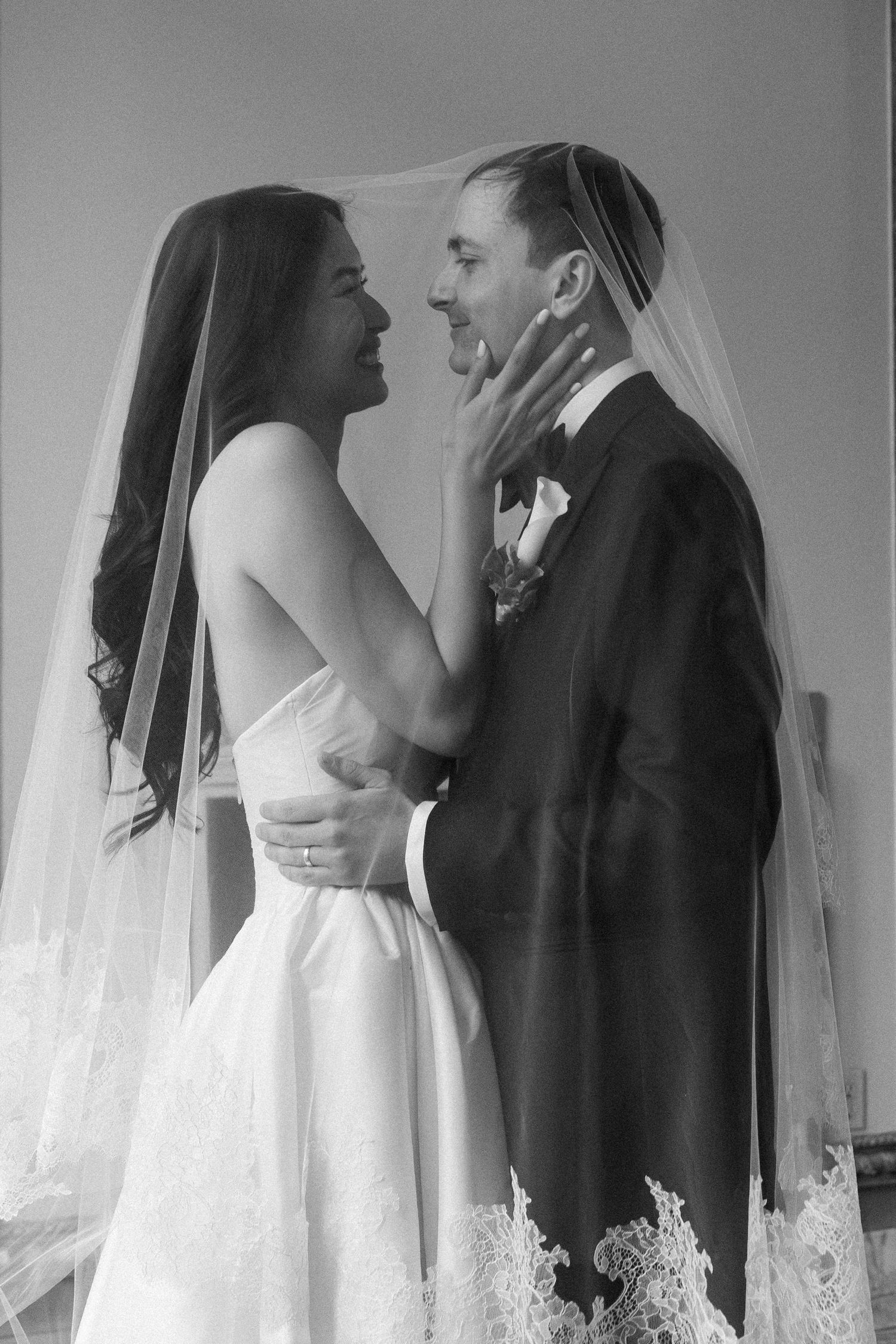 Black and white photo of bride and groom sharing a romantic moment under a lace-trimmed veil on their wedding day.