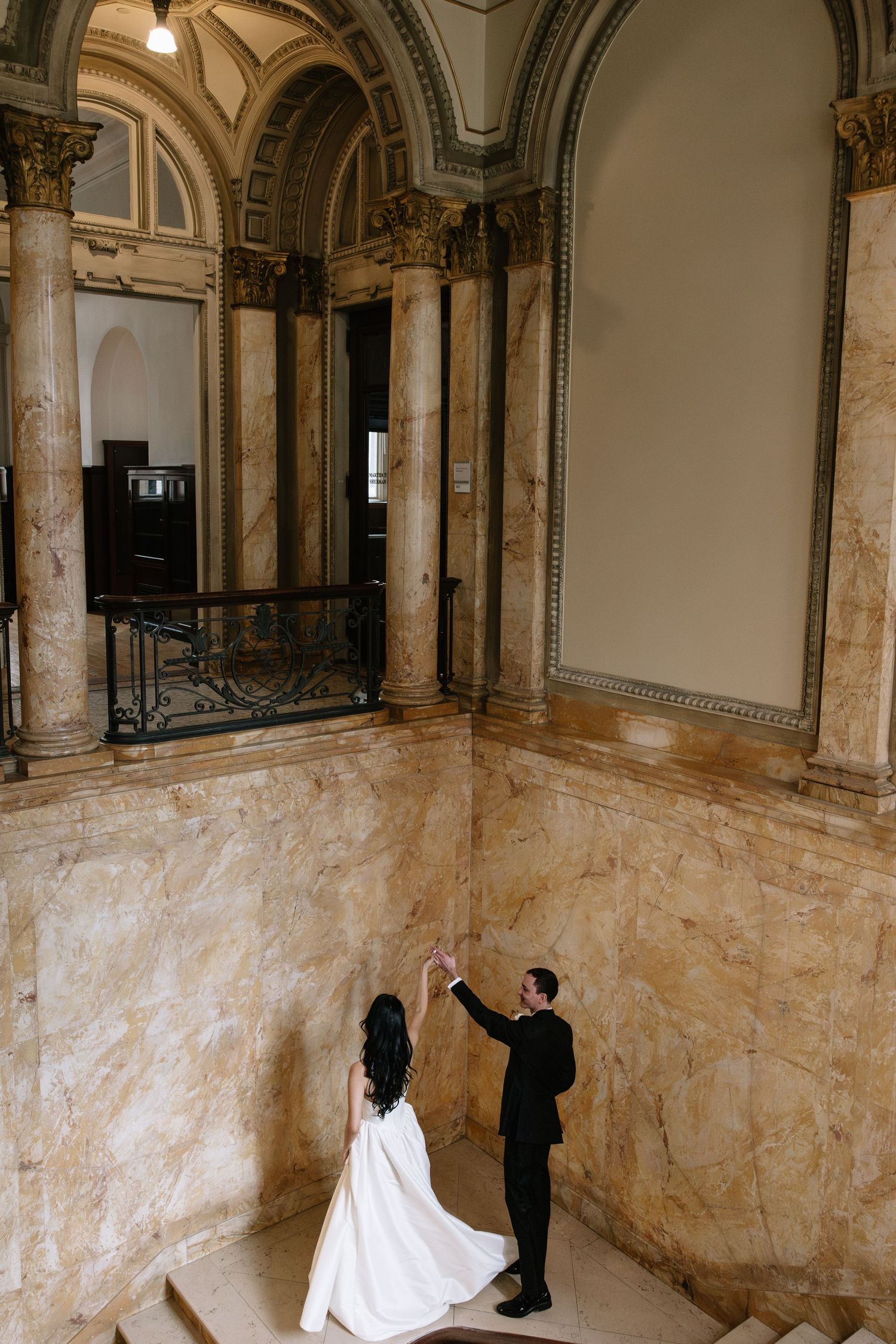 Bride and groom dance in grand marble staircase hall with ornate arched columns at elegant wedding venue.