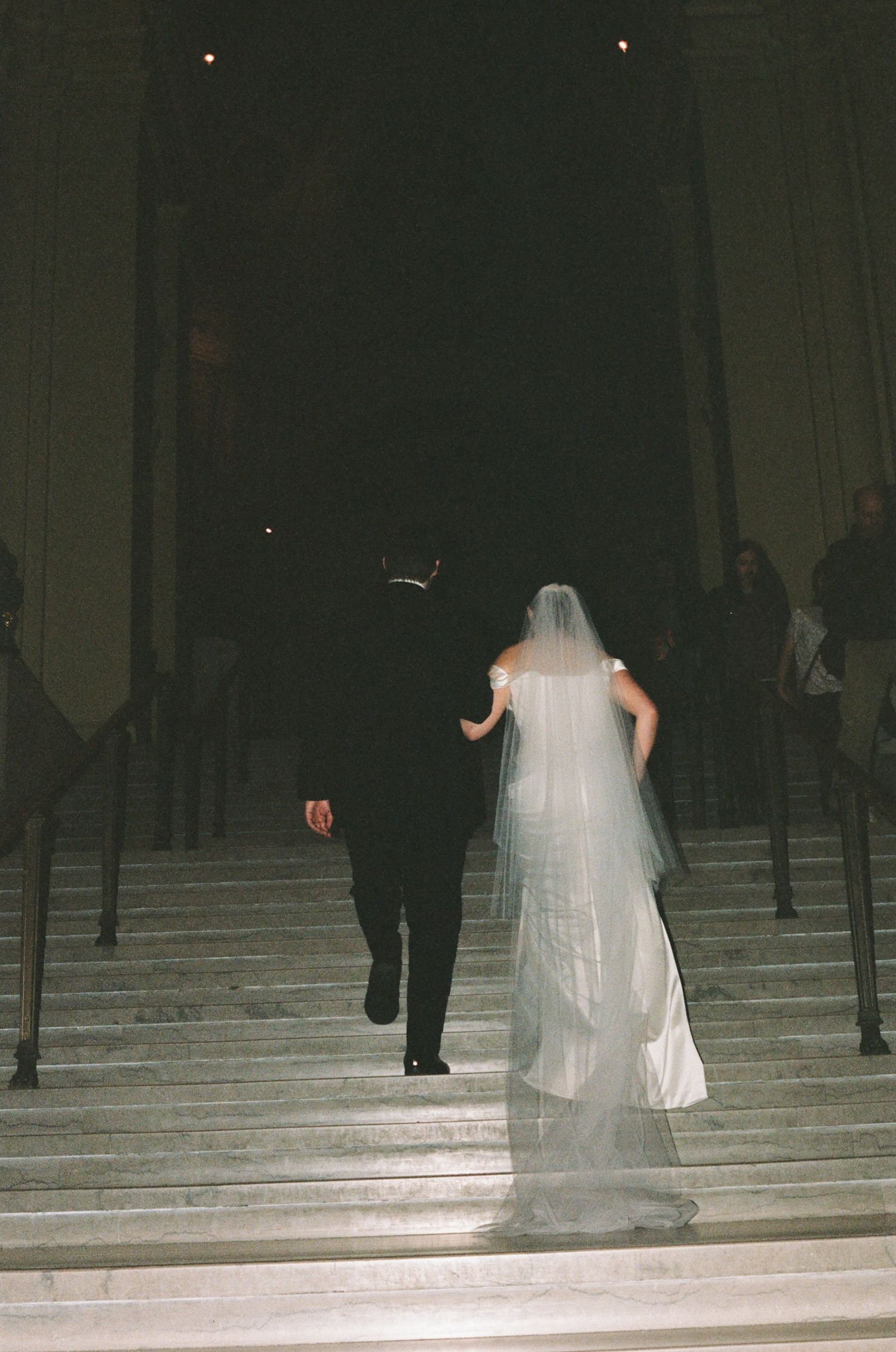 Bride and groom walking up grand stone stairs at night, woman in white wedding gown with veil, man in black tuxedo.