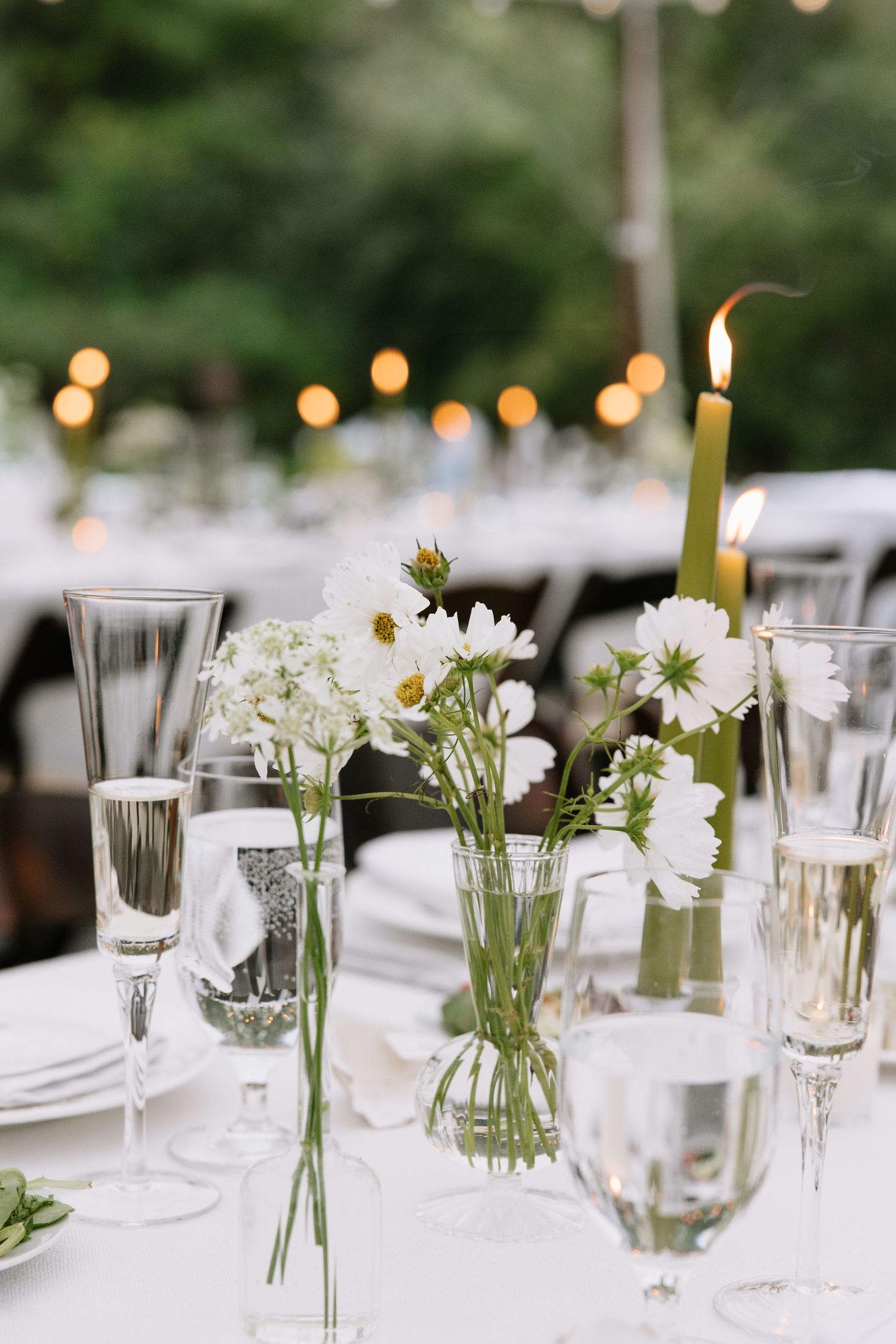 Elegant wedding reception table with white wildflowers, champagne flutes, and a green taper candle under string lights.
