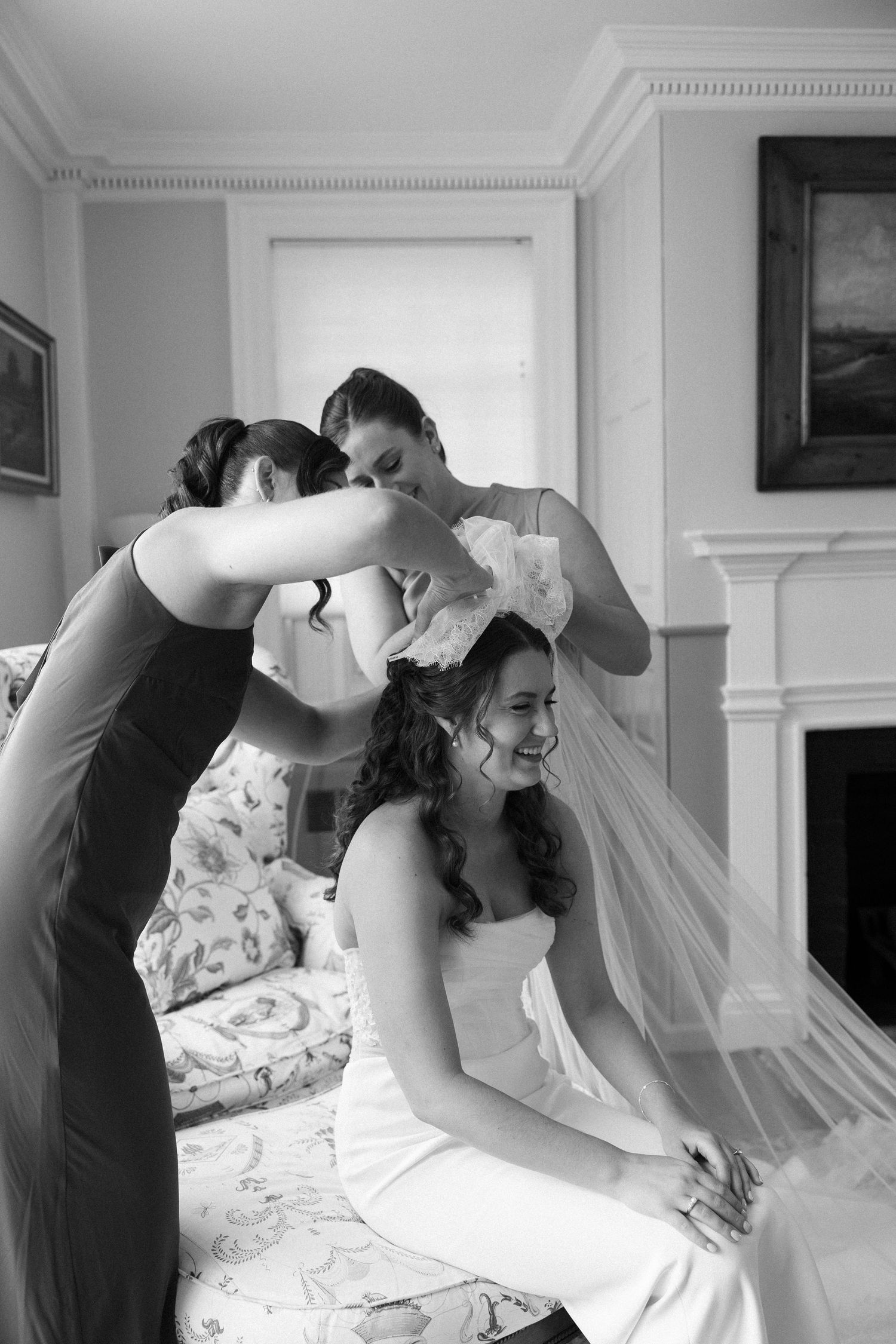 Bridesmaids helping a smiling bride put on her wedding veil in an elegant room, captured in black and white.
