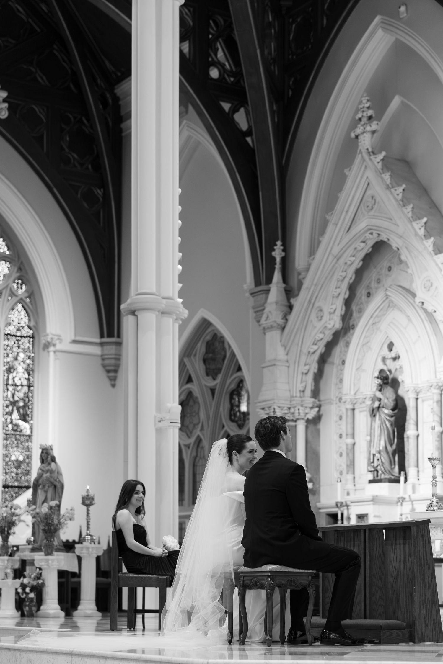 Bride and groom kneel at church altar during elegant black and white wedding ceremony with Gothic arched columns.