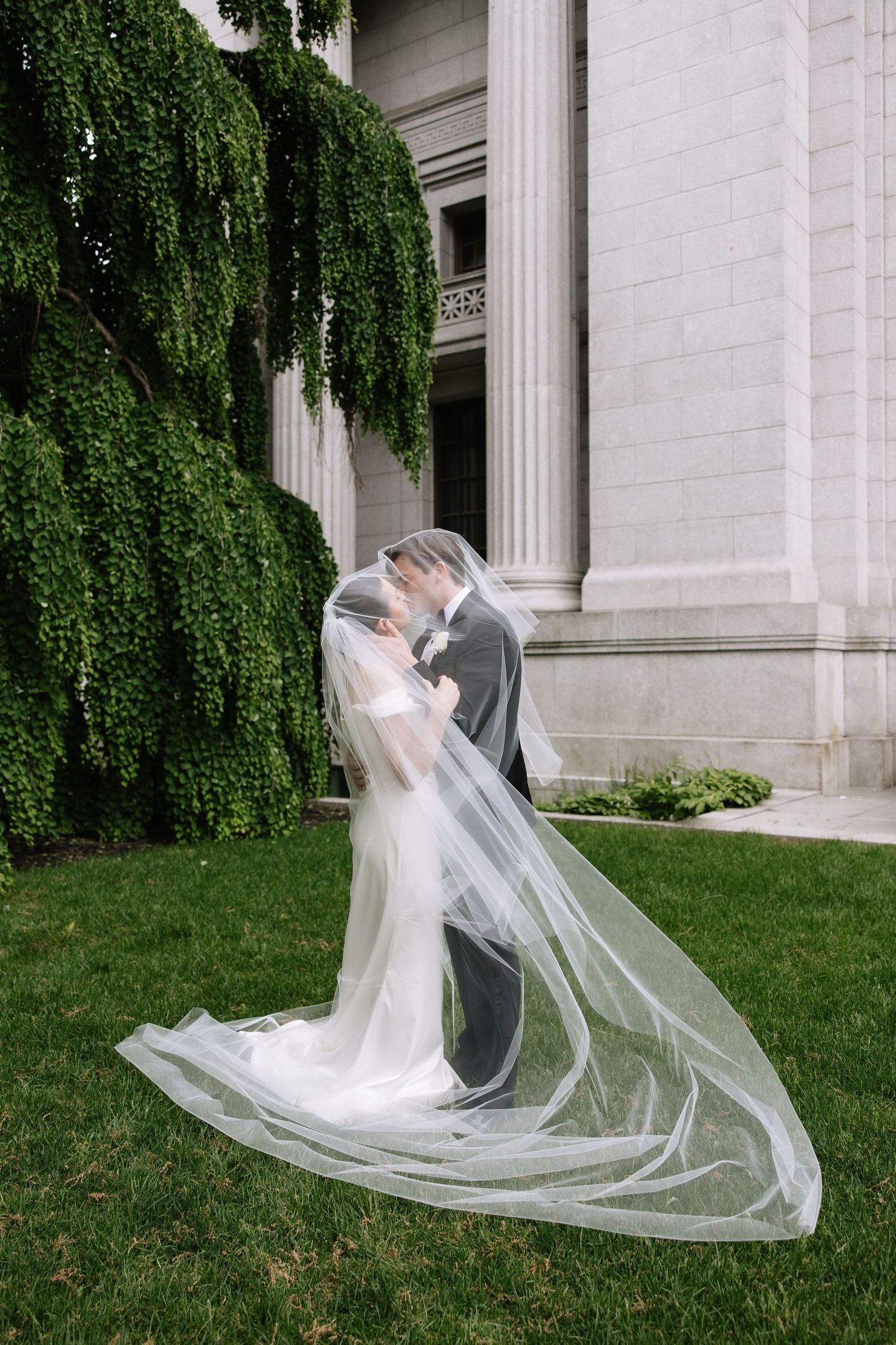 Bride and groom share a romantic kiss under a flowing cathedral veil on lush green lawn beside elegant stone building.