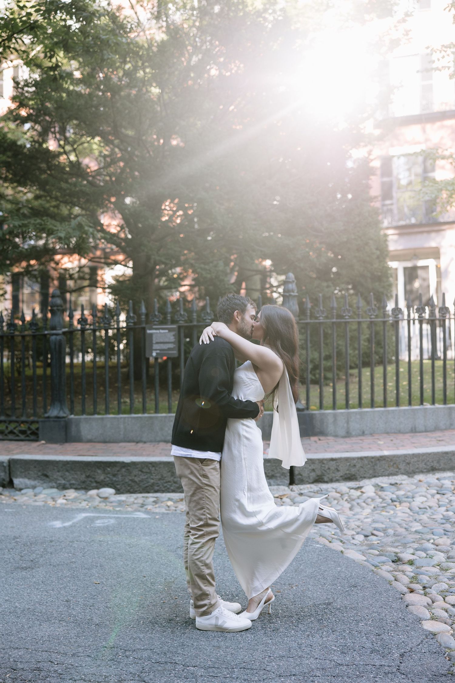 Romantic couple embracing on a cobblestone street with sunlight streaming through trees.