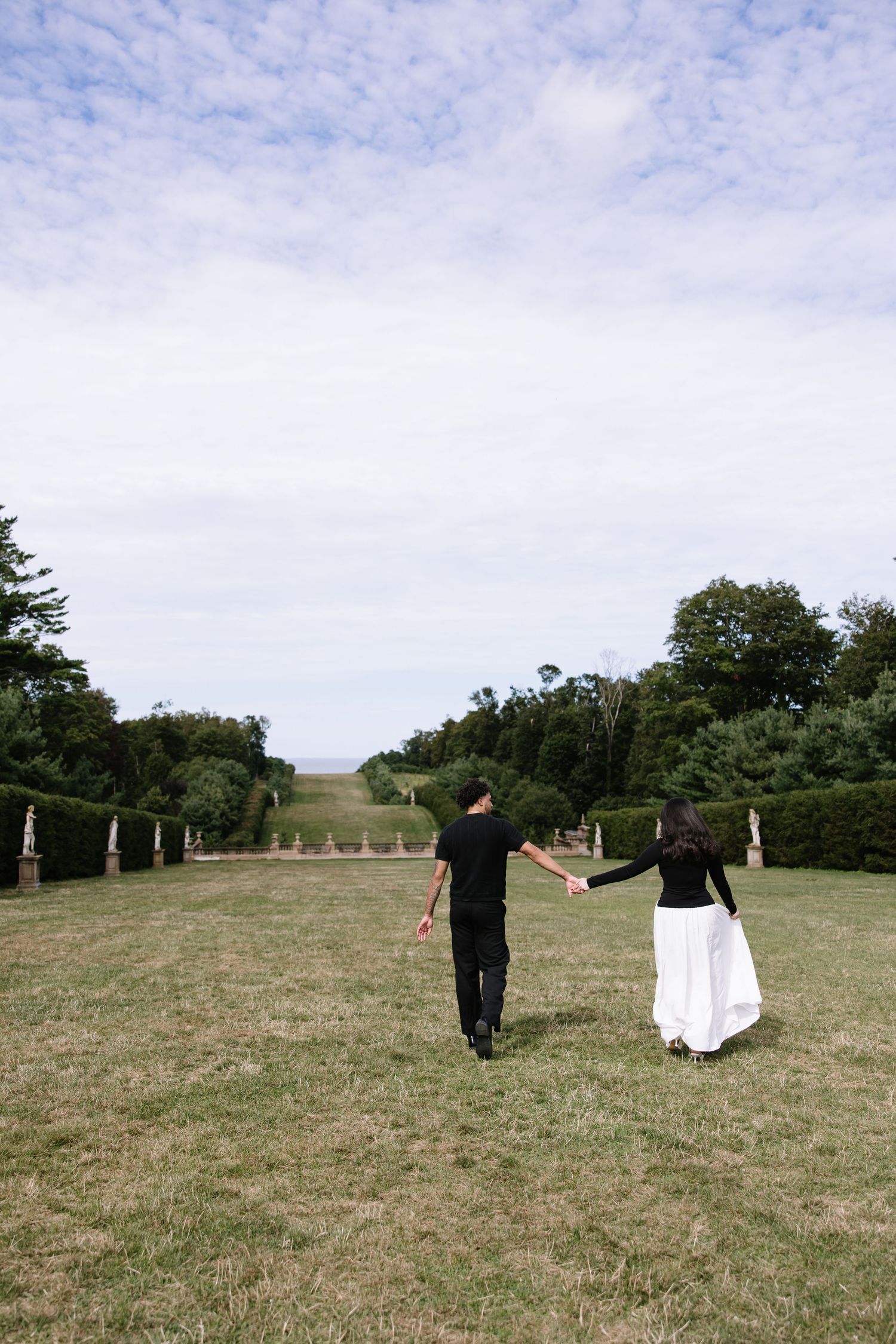 A couple walking hand in hand across a grassy field under cloudy skies.