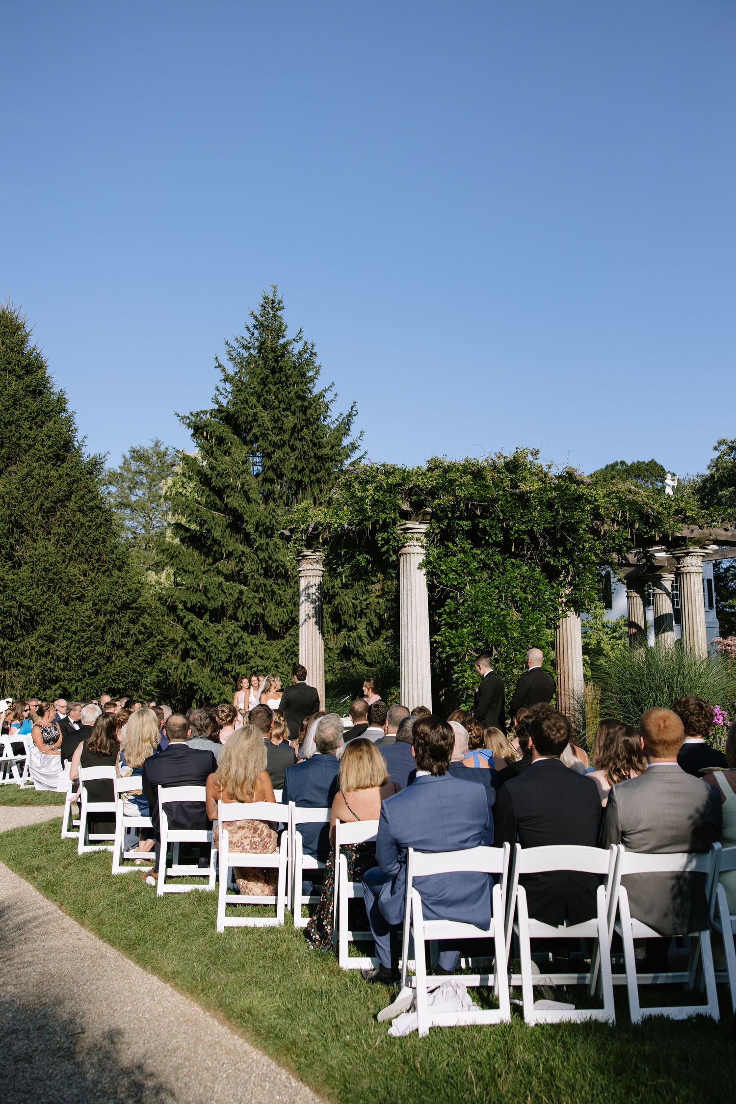 Outdoor garden wedding ceremony setup with white chairs and classical stone columns against a bright blue sky.