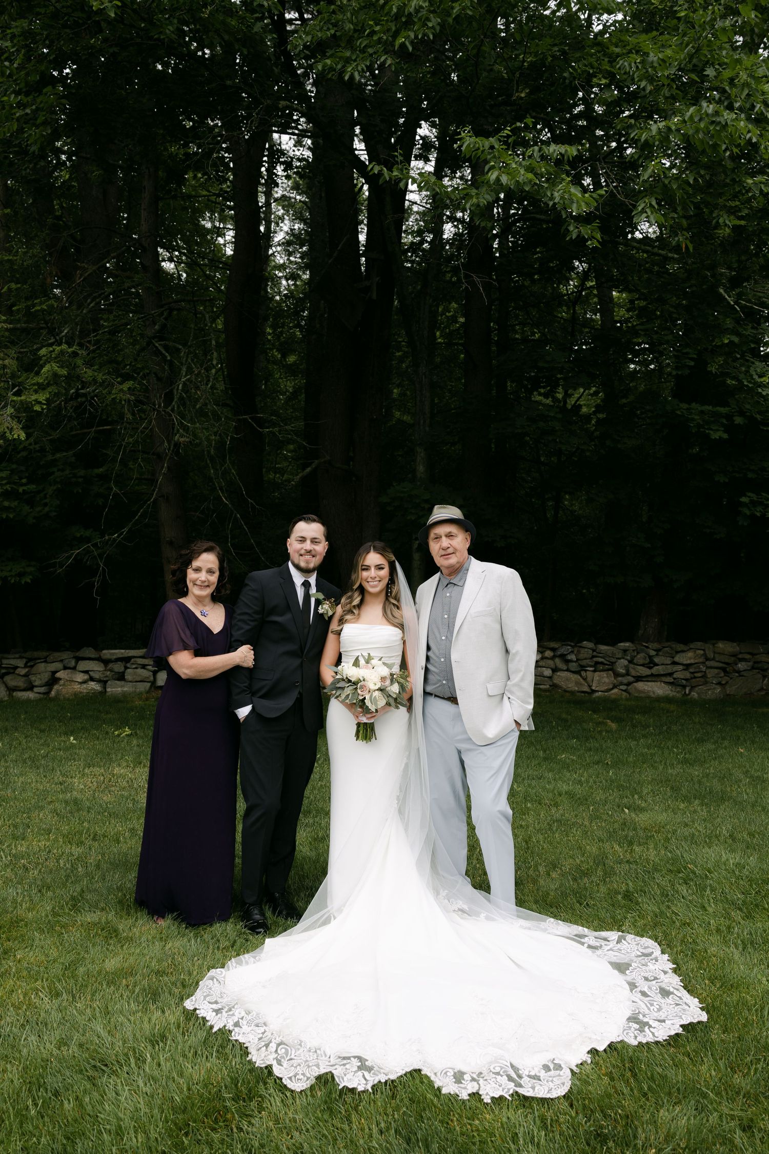 Wedding party poses on green lawn with stone wall and forest backdrop.