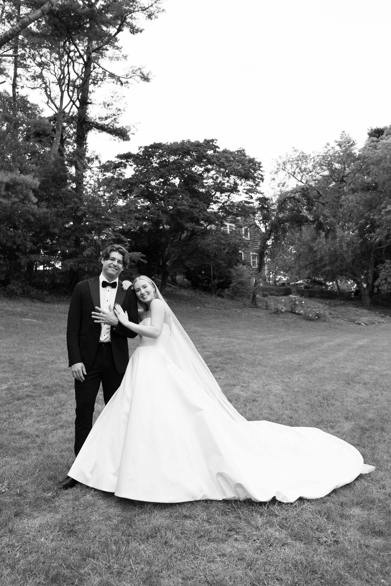 Black and white portrait of a couple in wedding attire posing on a grassy lawn.