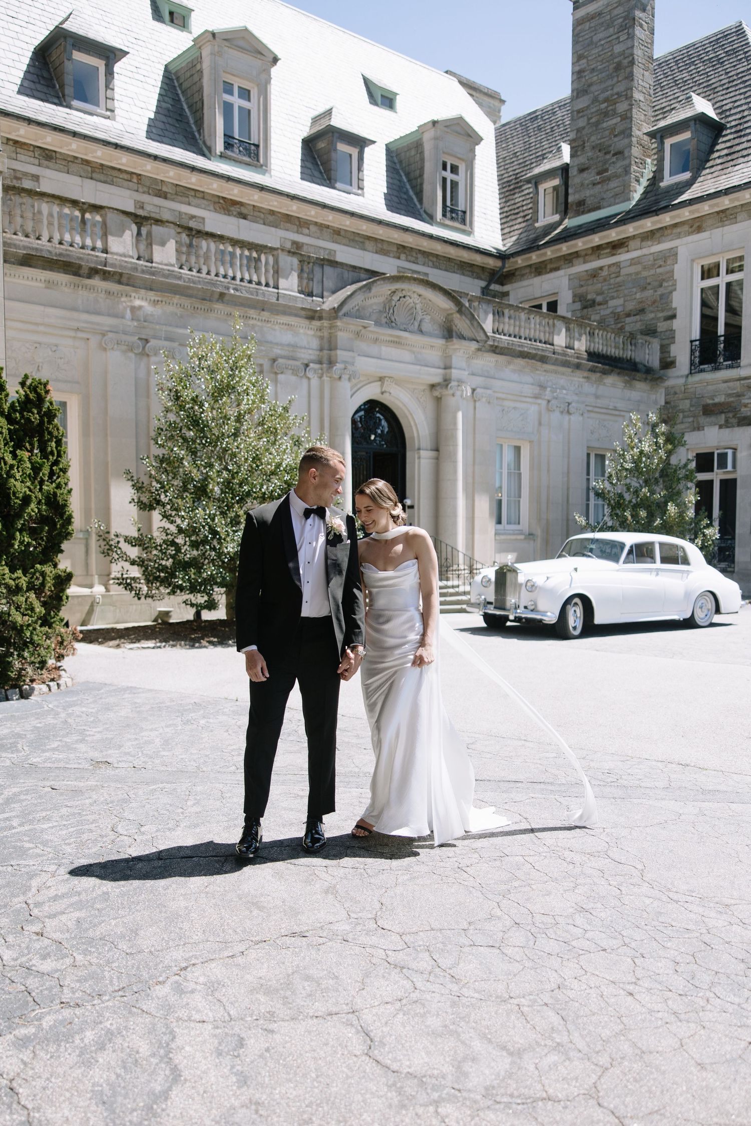 Bride and groom stand in front of elegant stone mansion with vintage white Rolls-Royce, bride in satin gown, groom in black tuxedo.