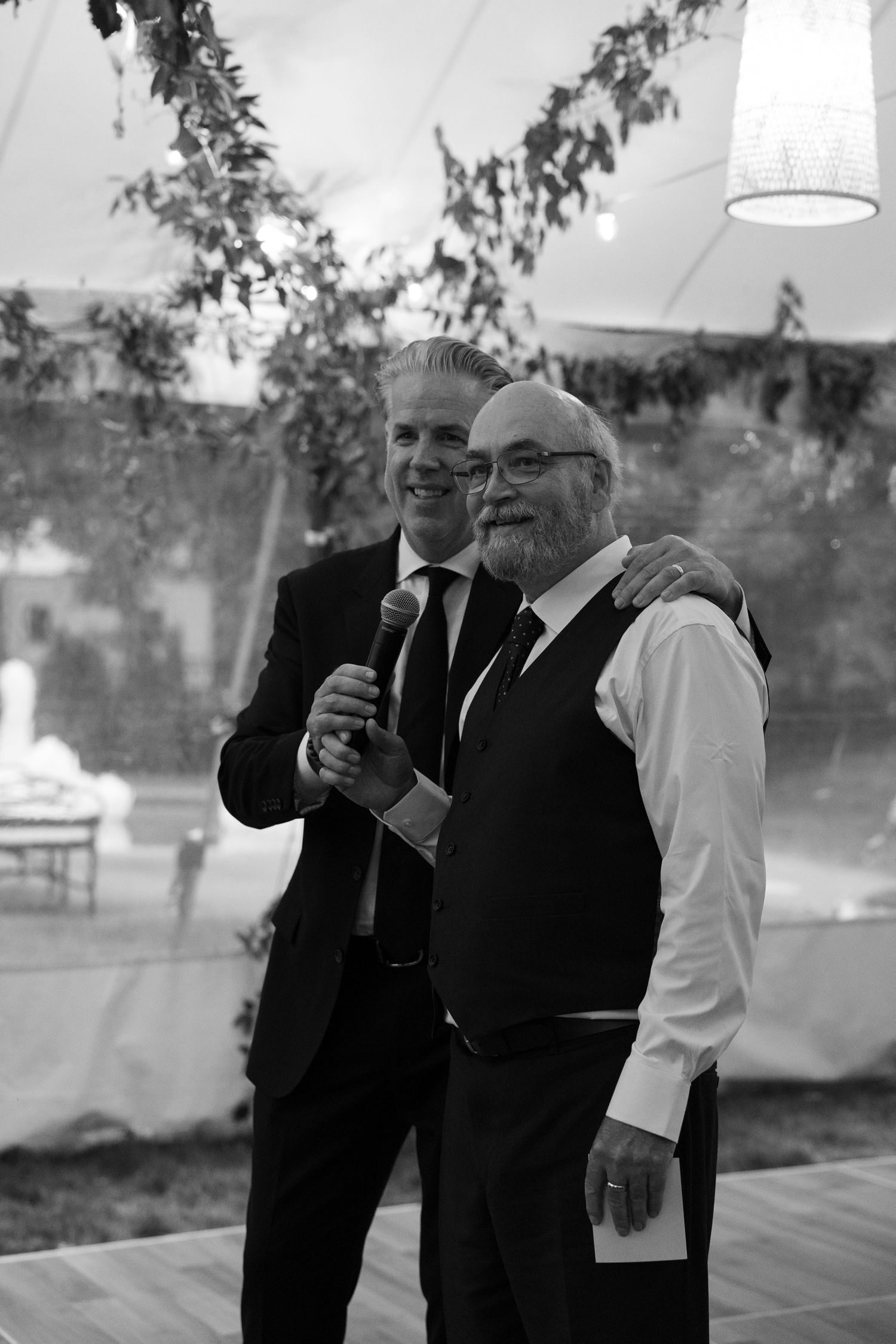 Two men in formal suits share a warm embrace while one holds a microphone at an elegant wedding reception under a tent.