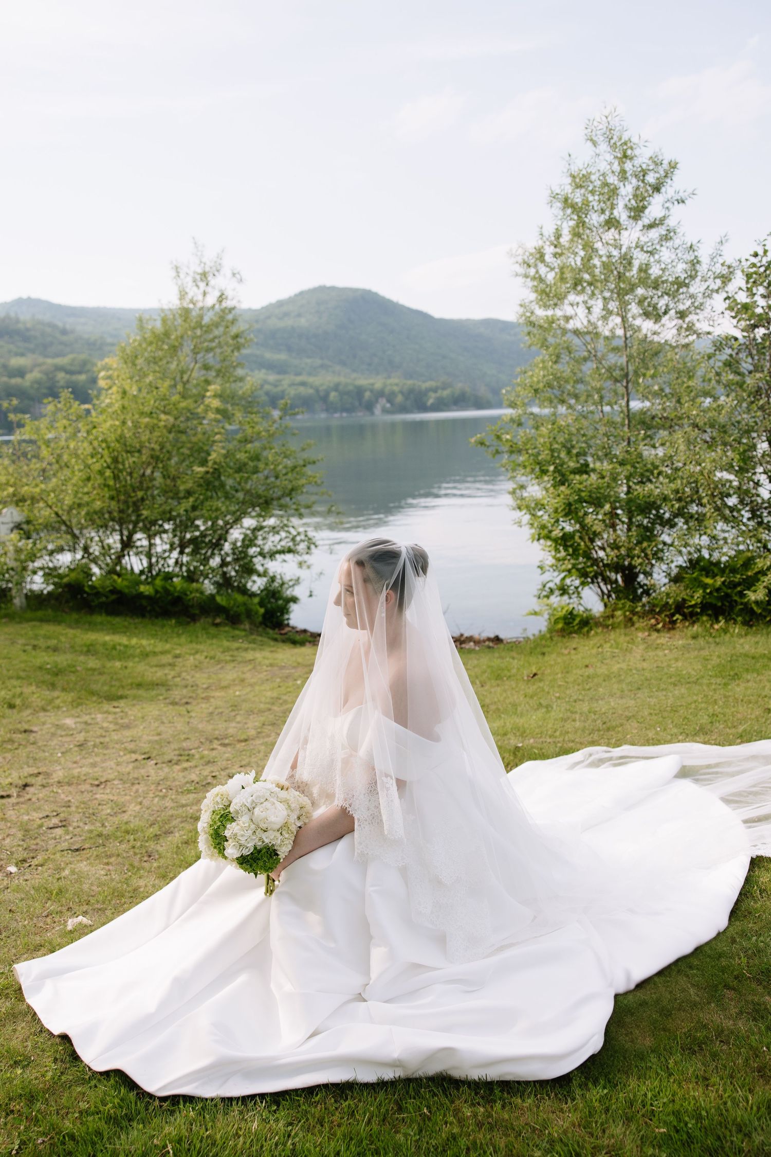 A bride in a white gown and veil sits on grass holding a bouquet with a scenic lake and mountains behind her.