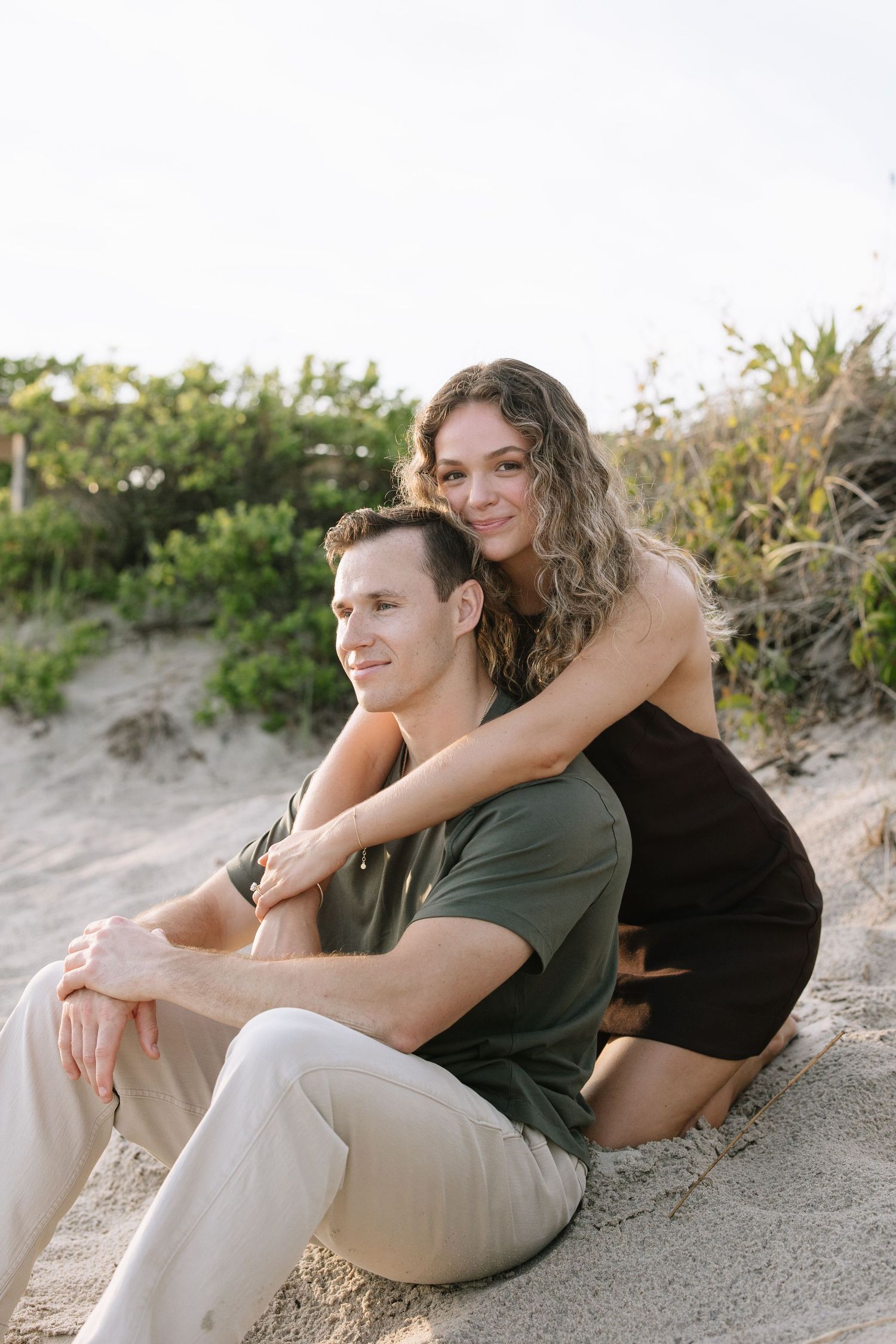 Couple sitting together on sandy beach dunes surrounded by coastal greenery at golden hour.