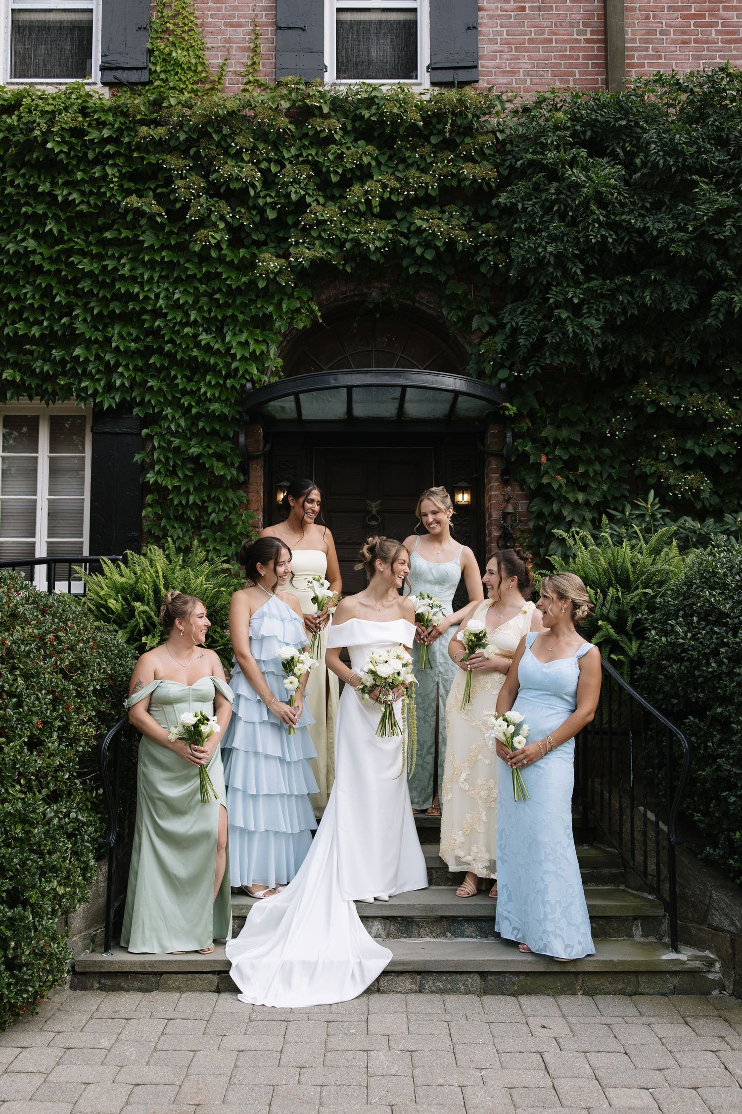 Bride in white gown poses with six bridesmaids in mismatched blue and green dresses on ivy-covered brick building steps.