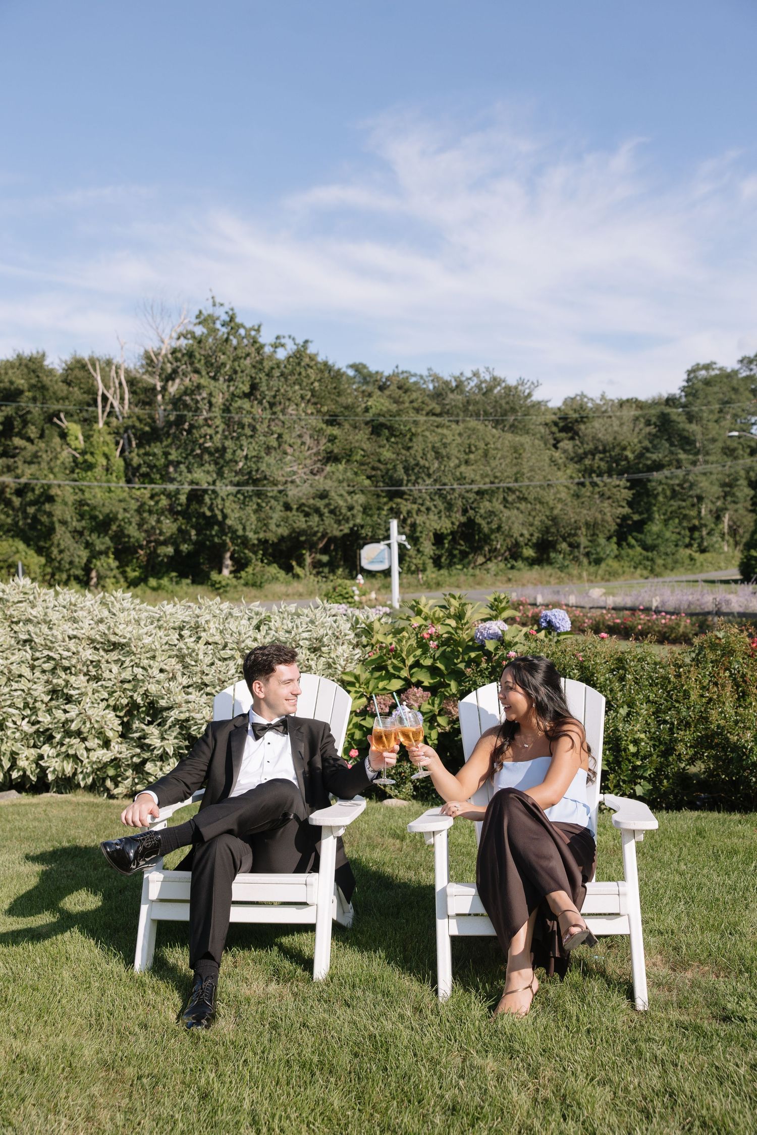 Couple in formal attire toasting with champagne glasses while seated in white Adirondack chairs on lawn.