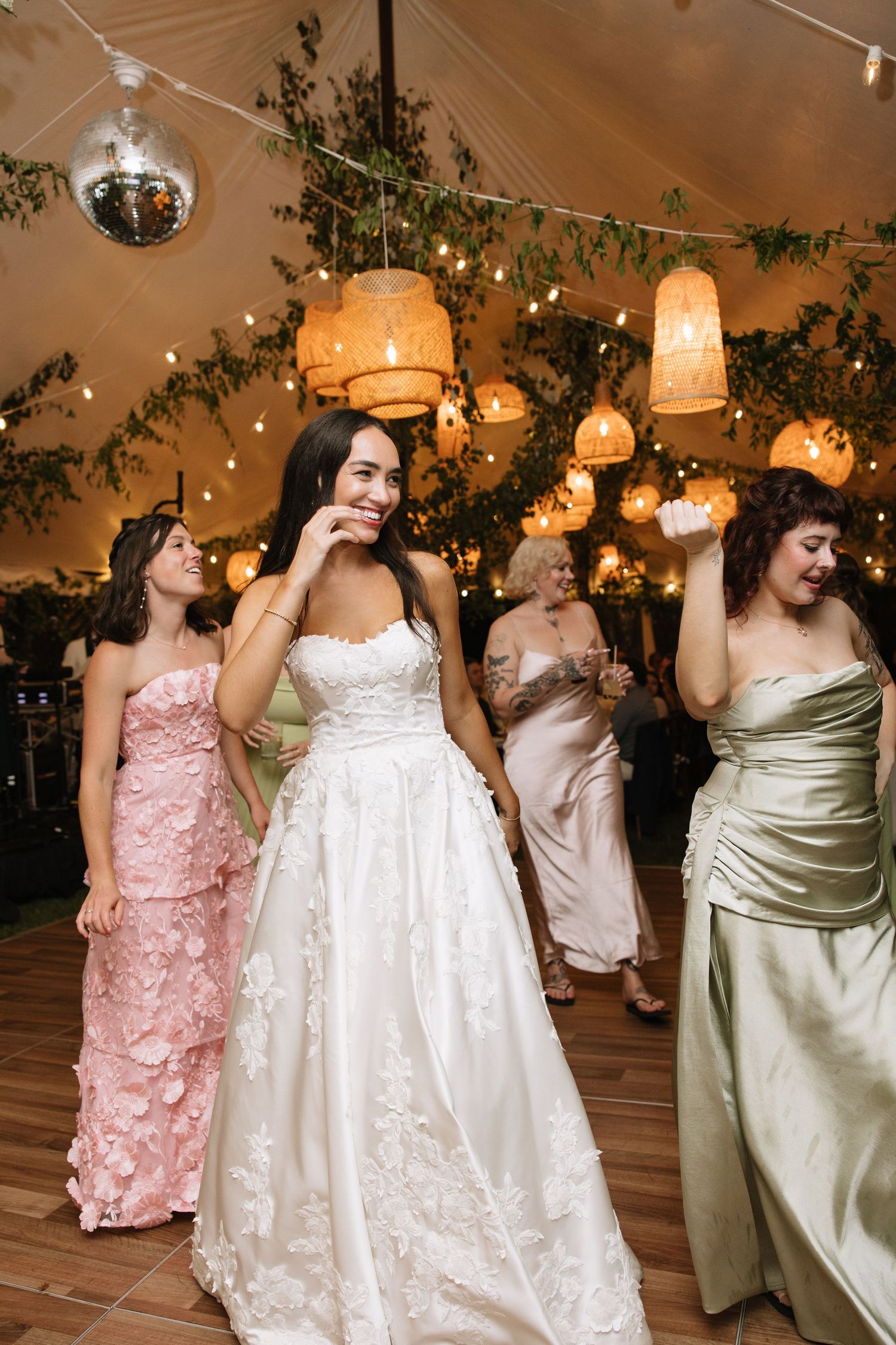Bride in a floral lace ballgown dancing joyfully with guests under warm lights and hanging lanterns at wedding reception.