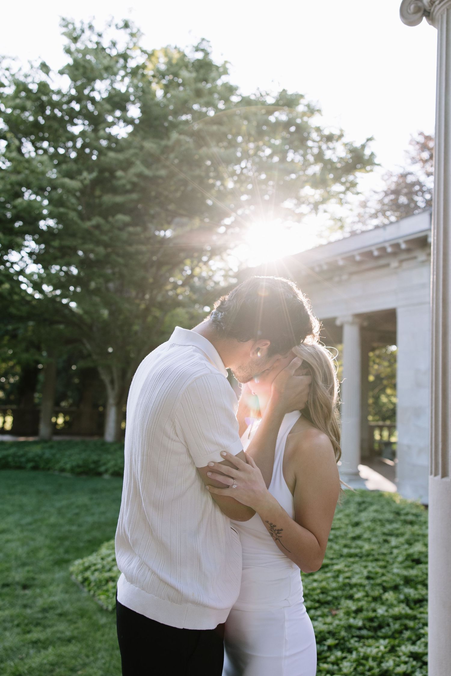 Couple kissing in golden sunlight outdoors, wearing white outfits, surrounded by lush green trees and a classic white building.