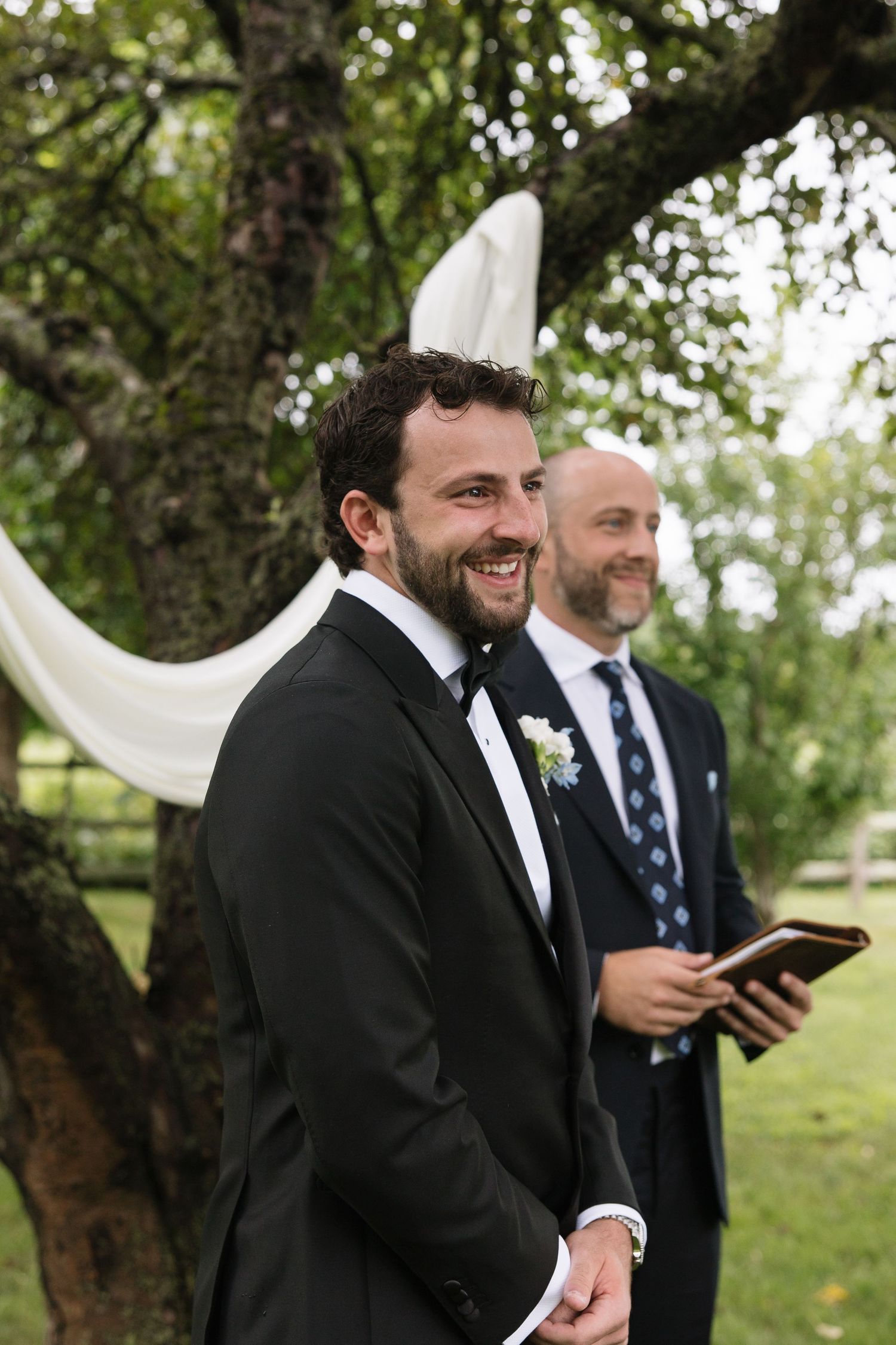 Smiling groom in black tuxedo with boutonniere stands at outdoor wedding ceremony beneath draped tree.
