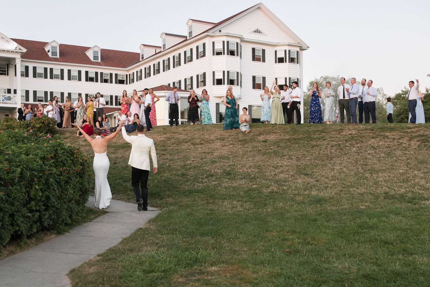 Wedding couple walks toward guests gathered on lawn outside elegant white colonial venue at outdoor reception.