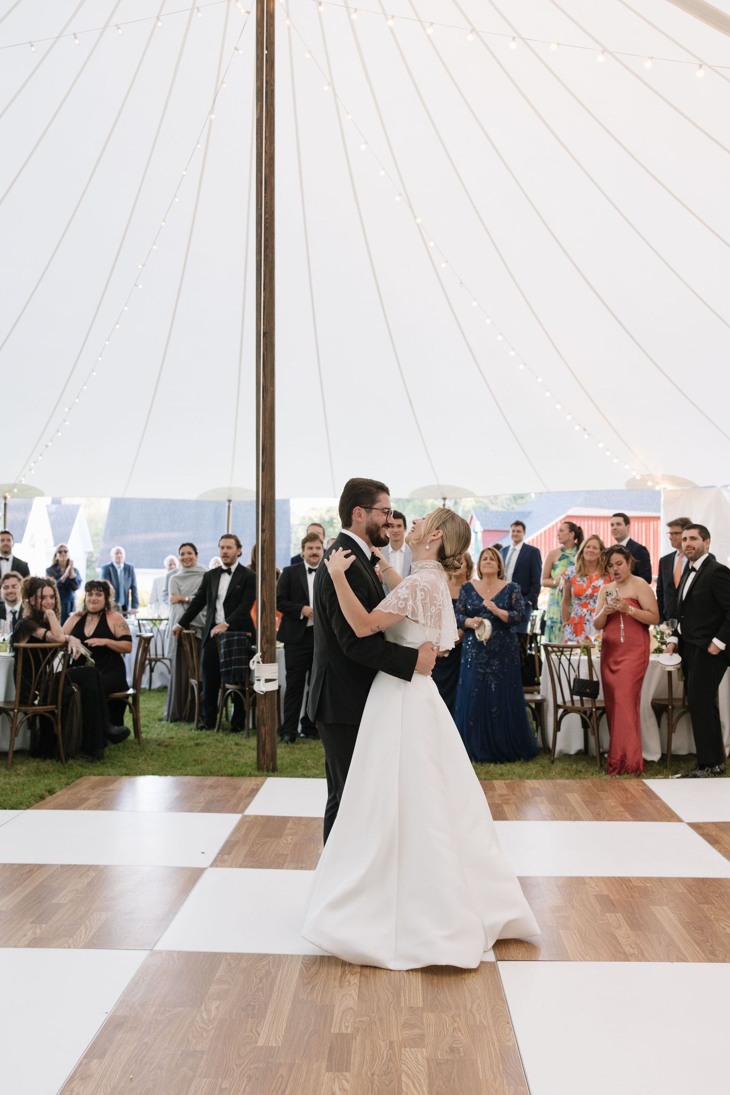 Bride and groom share their first dance on checkered floor under a white wedding tent with guests watching.