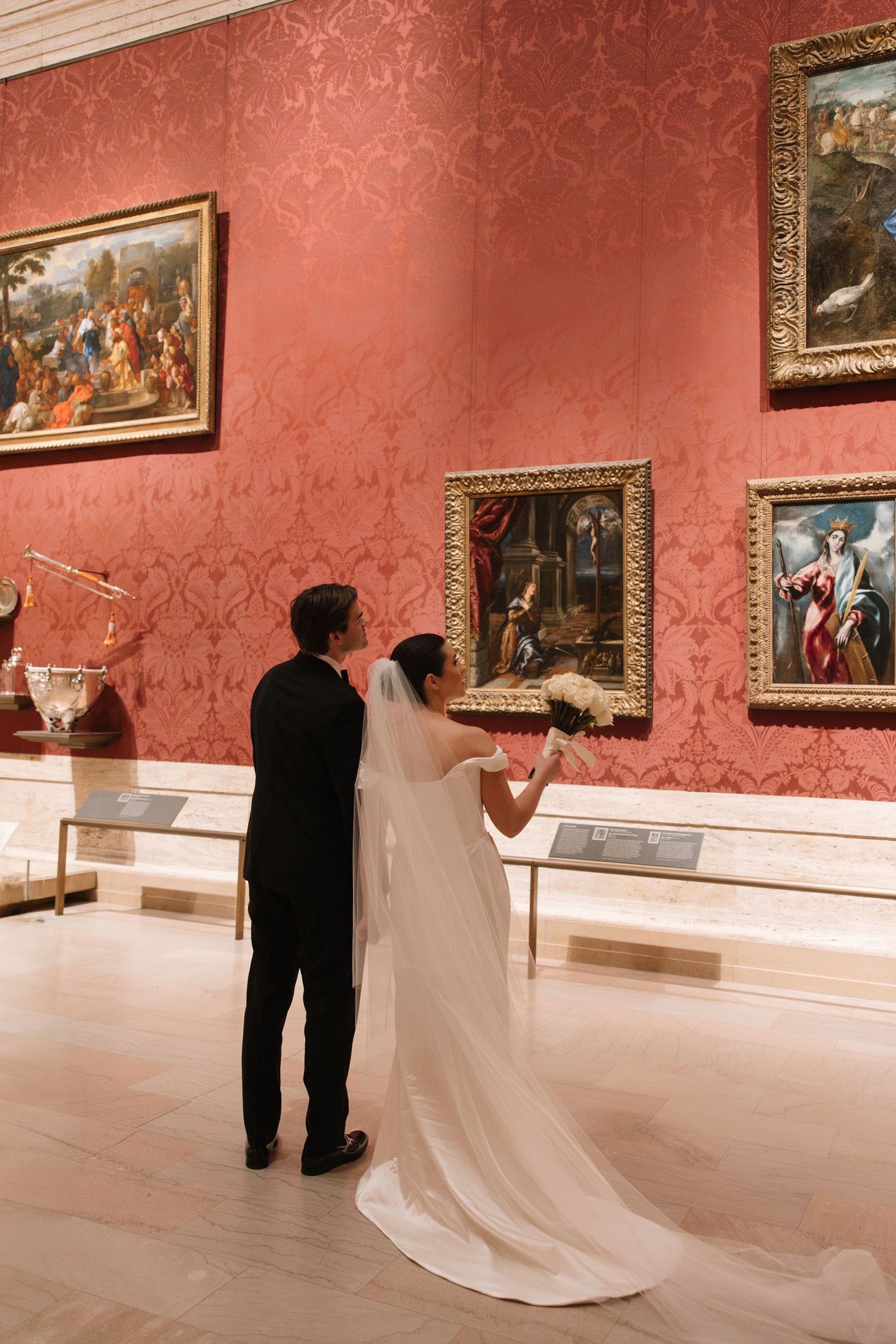 Bride and groom viewing classic paintings in ornate red gallery during wedding day museum visit.
