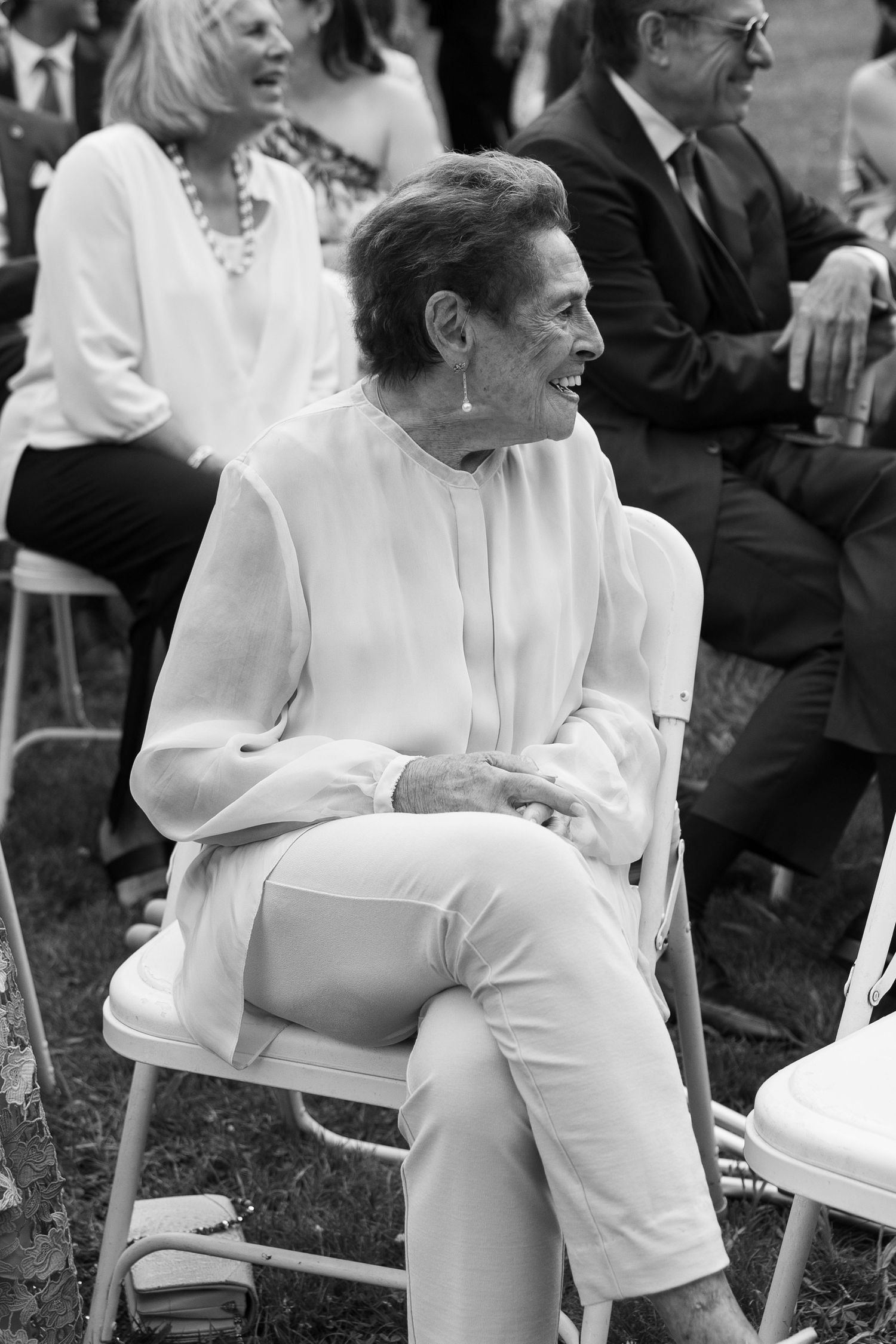 Elderly woman in white outfit laughing joyfully while seated at an outdoor wedding ceremony.