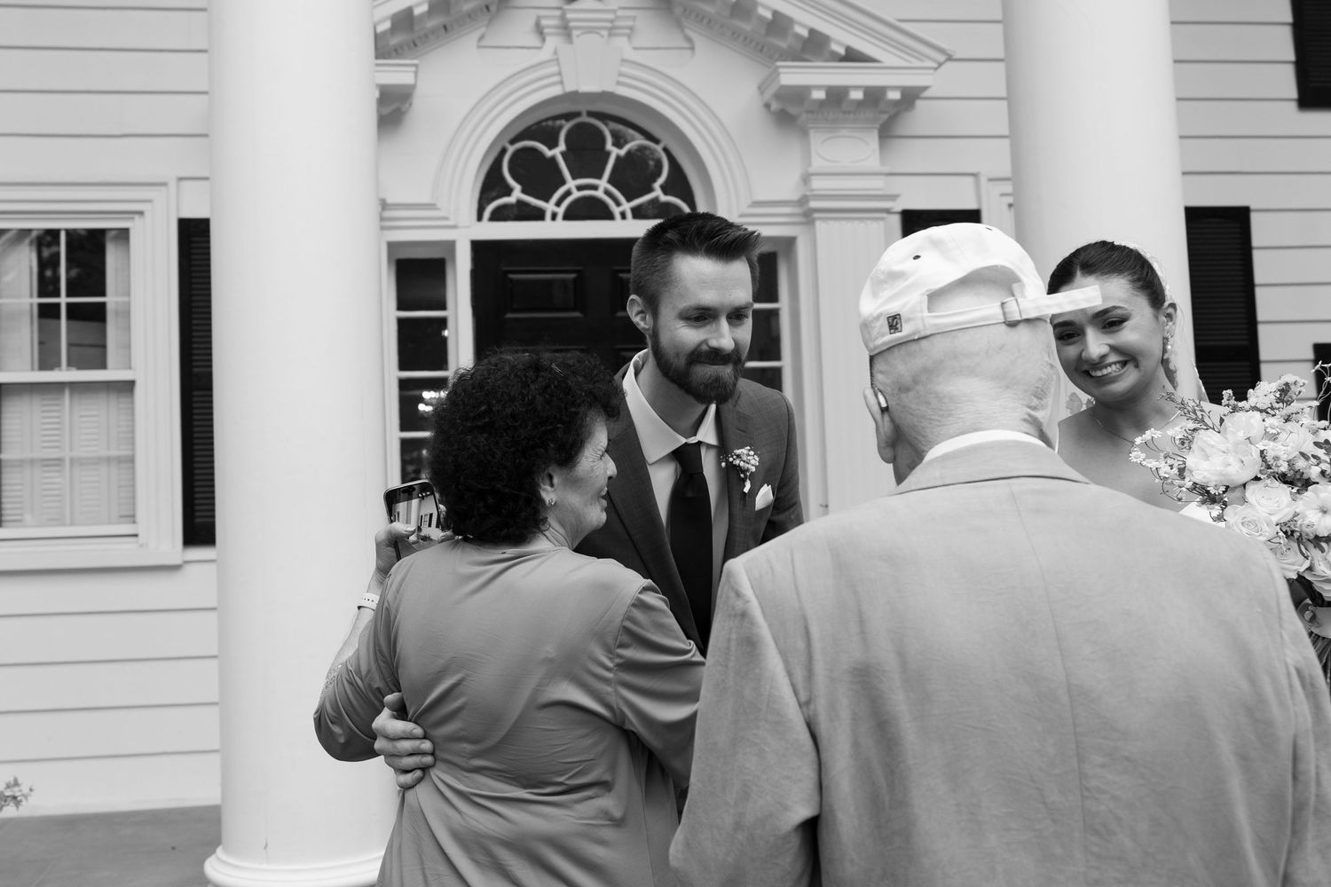Black and white photo of a groom greeting elderly guests outside a historic white colonial building at a wedding.