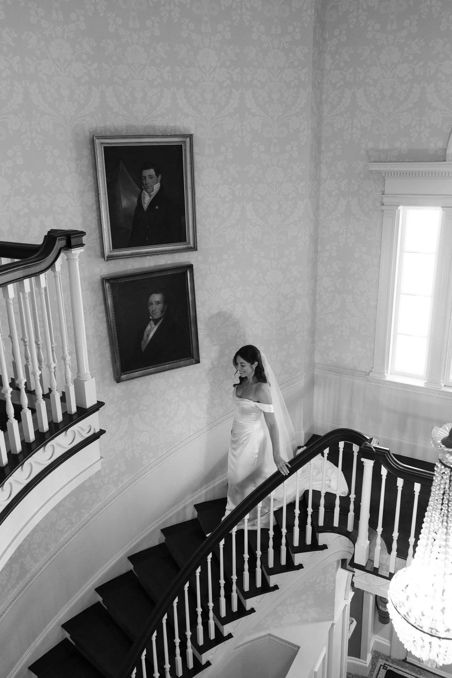 Bride in white gown and veil descends grand staircase in elegant venue with portrait paintings on wall.