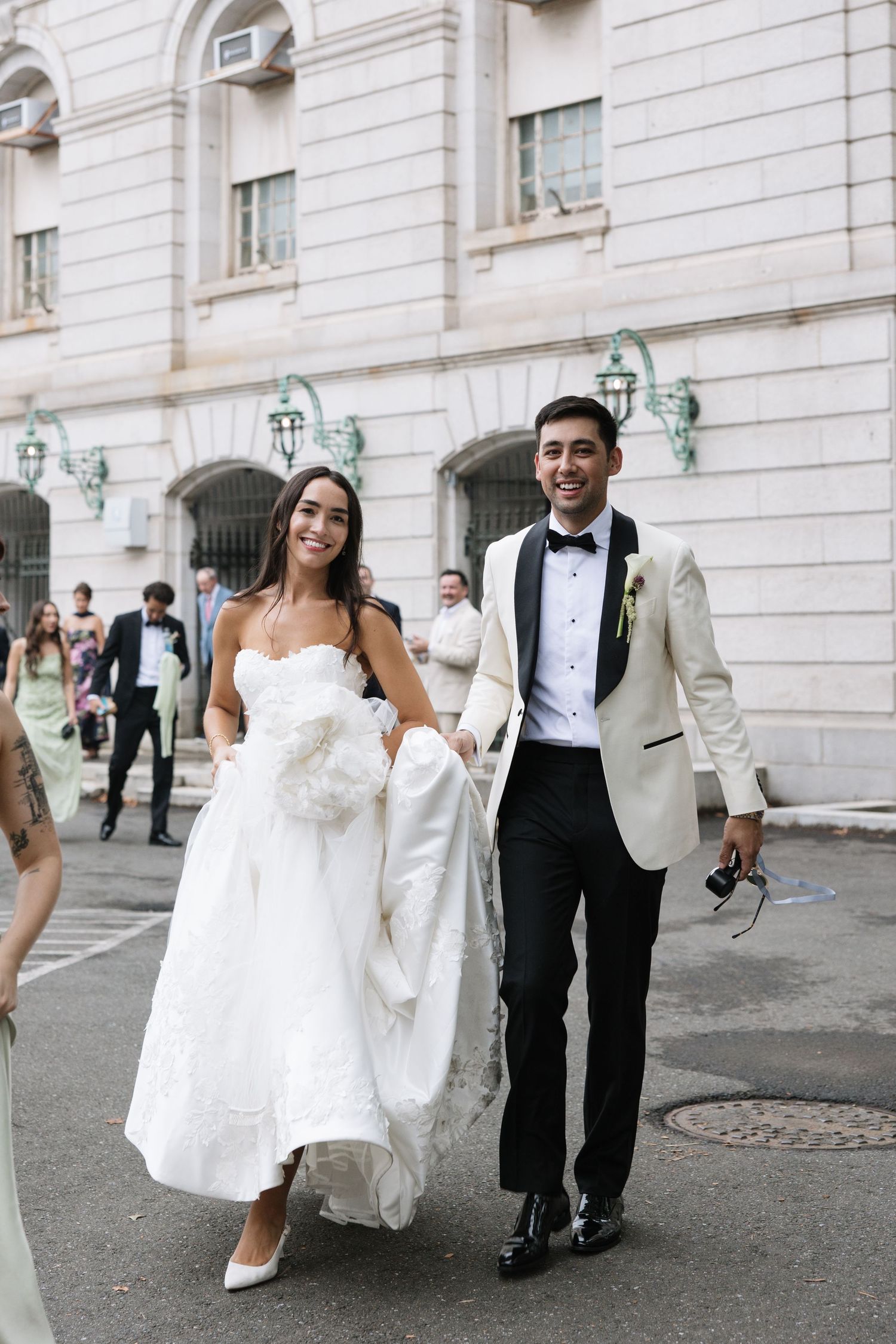 Smiling bride and groom in white tuxedo jacket walk hand-in-hand outside elegant building after ceremony.