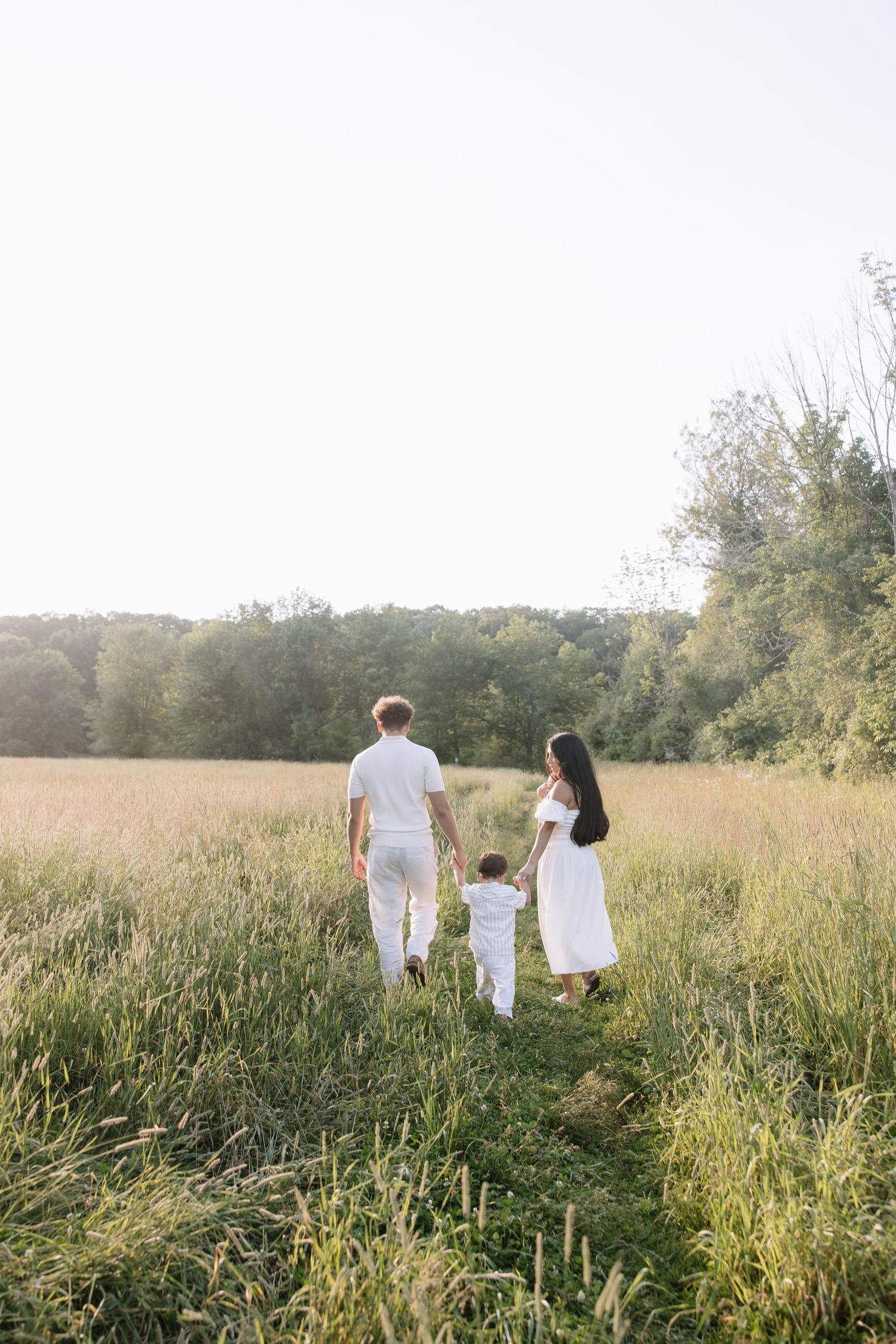 A family in white outfits walks hand-in-hand through a sunny green meadow with trees in the background.