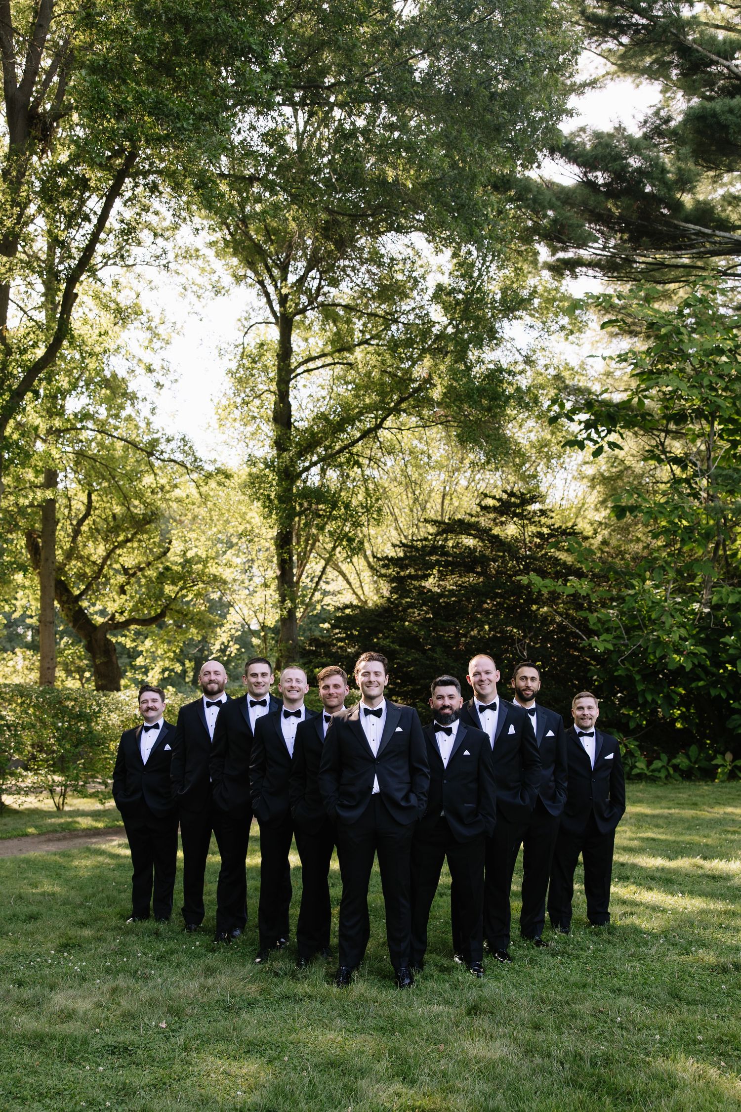 Groomsmen in black tuxedos pose for formal group photo on garden lawn.