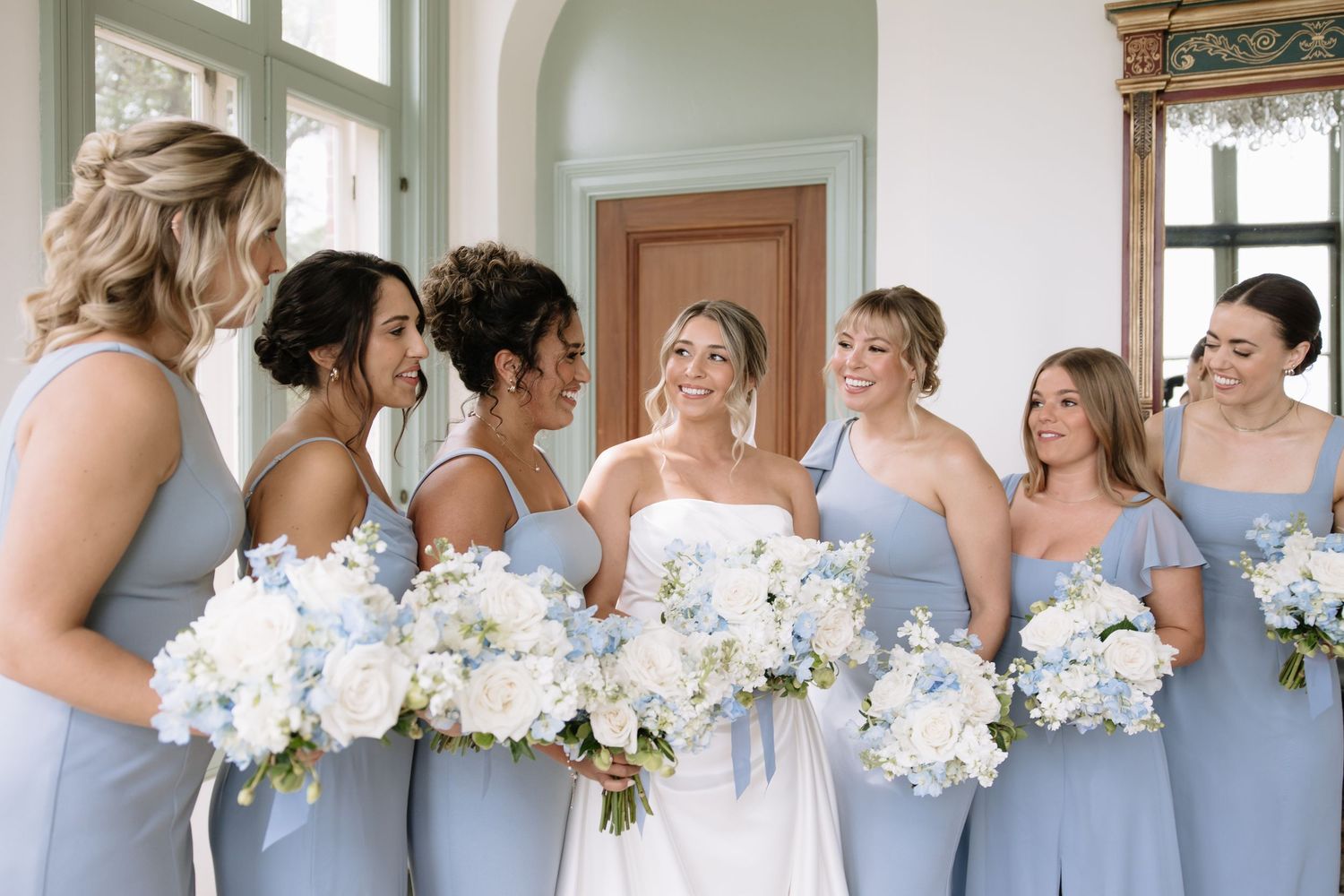 Wedding party with white bouquets gathered in elegant room with arched doorways.