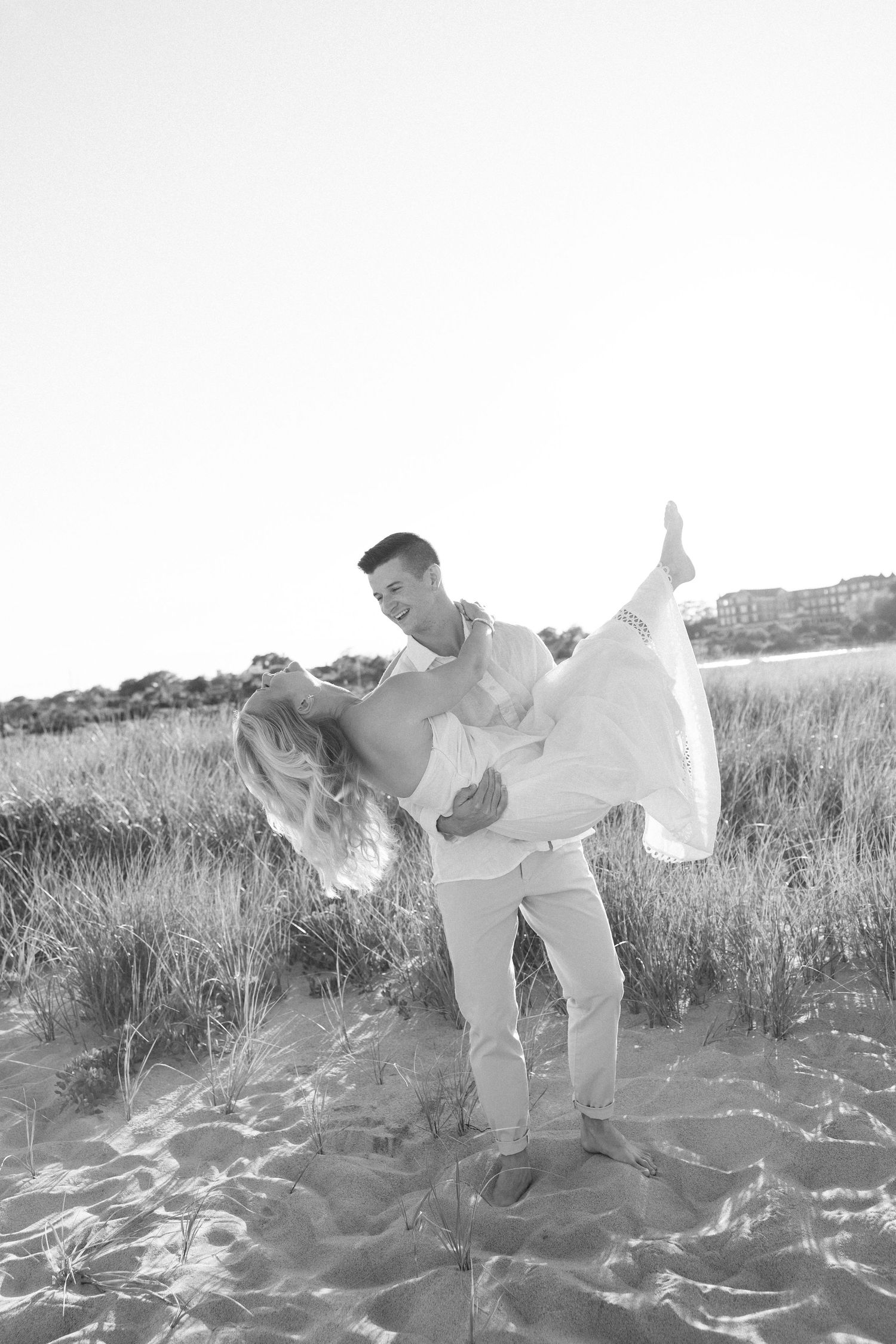 Black and white photo of a man lifting a woman on a sandy beach with grass dunes in the background.